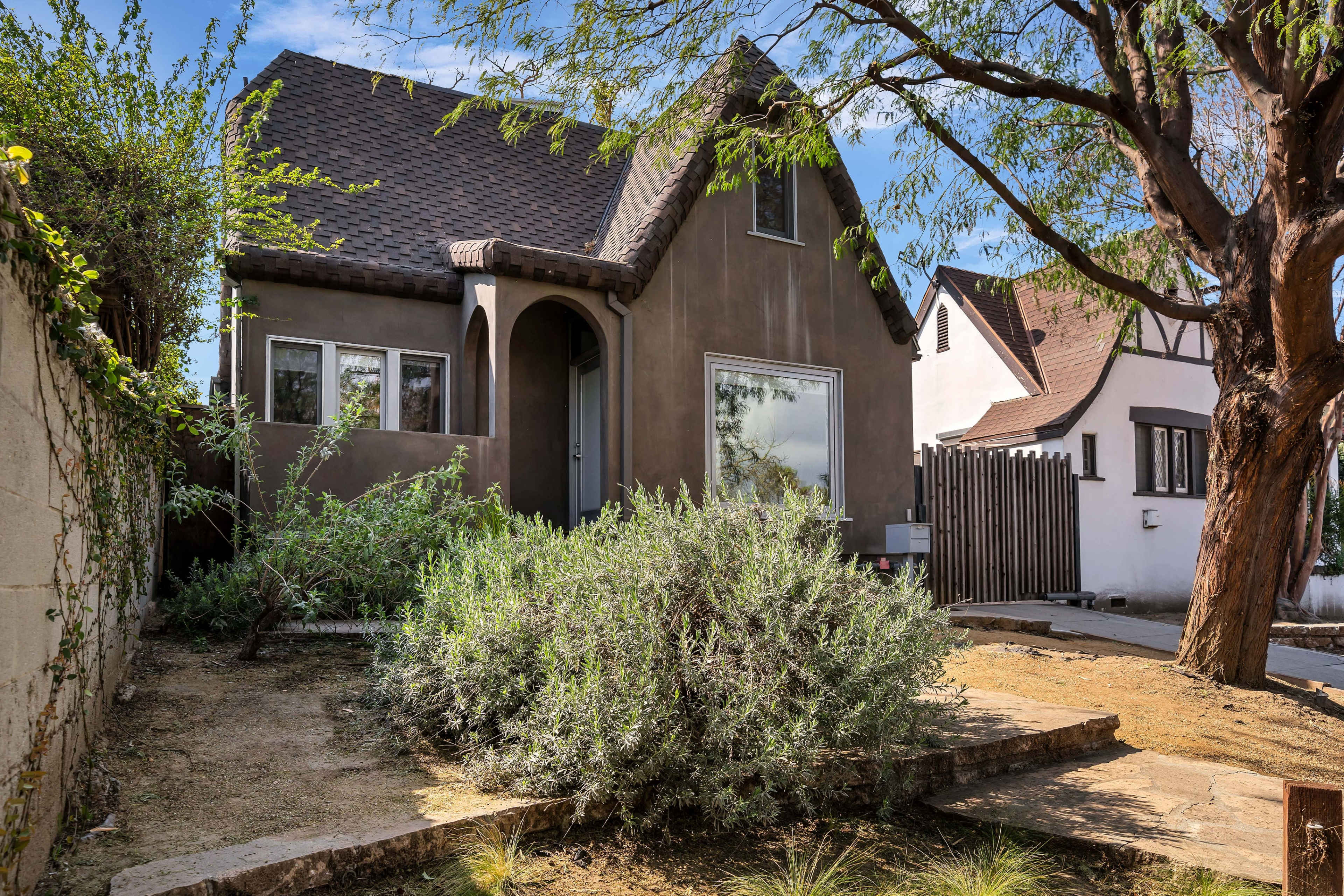 A small, two-story house with a brown exterior and a steep roof is situated in a landscaped yard surrounded by low-growing shrubs and trees.