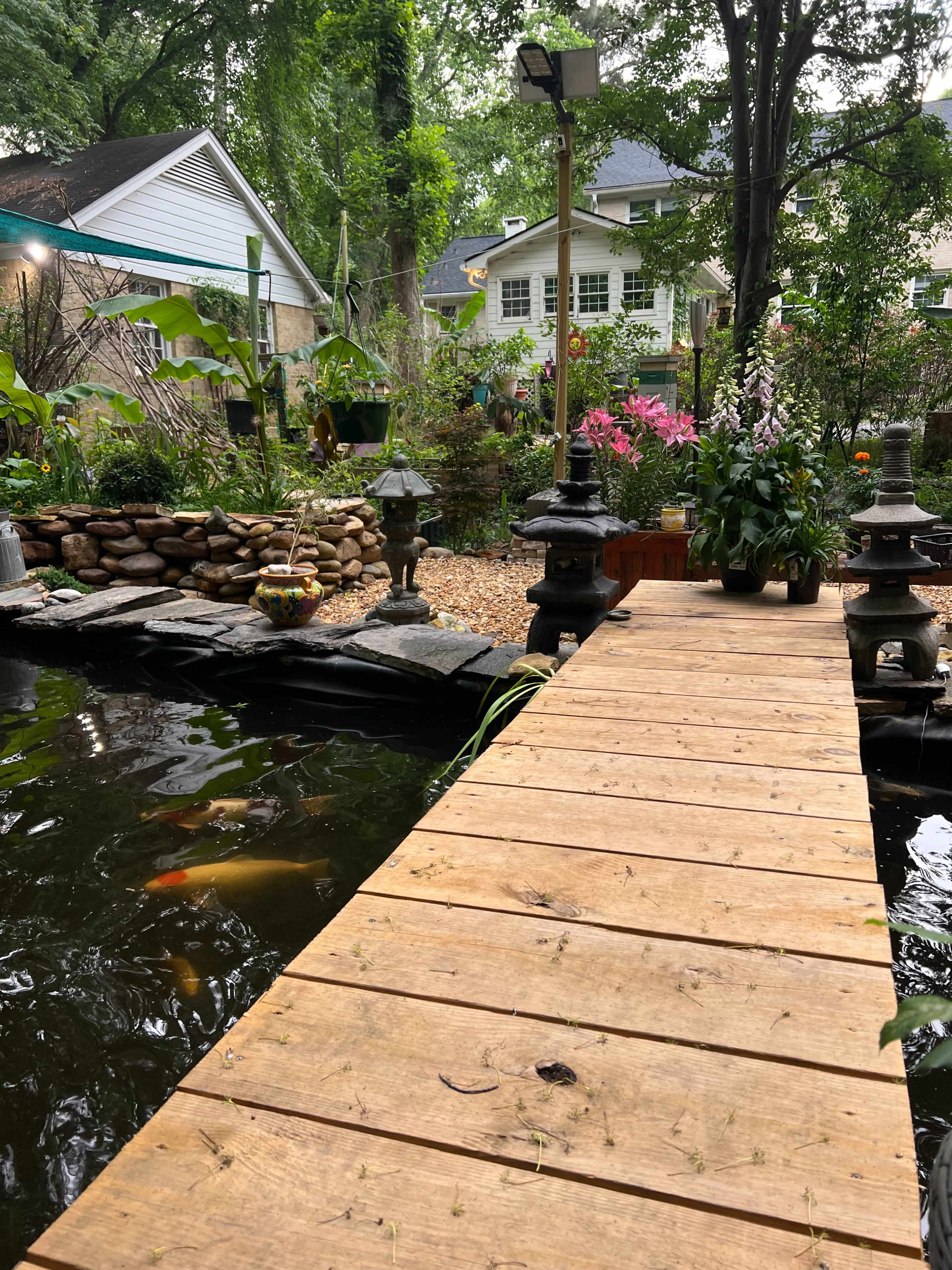 A wooden pathway extends over a pond surrounded by lush greenery and vibrant flowers, leading to a decorative stone wall and lanterns.