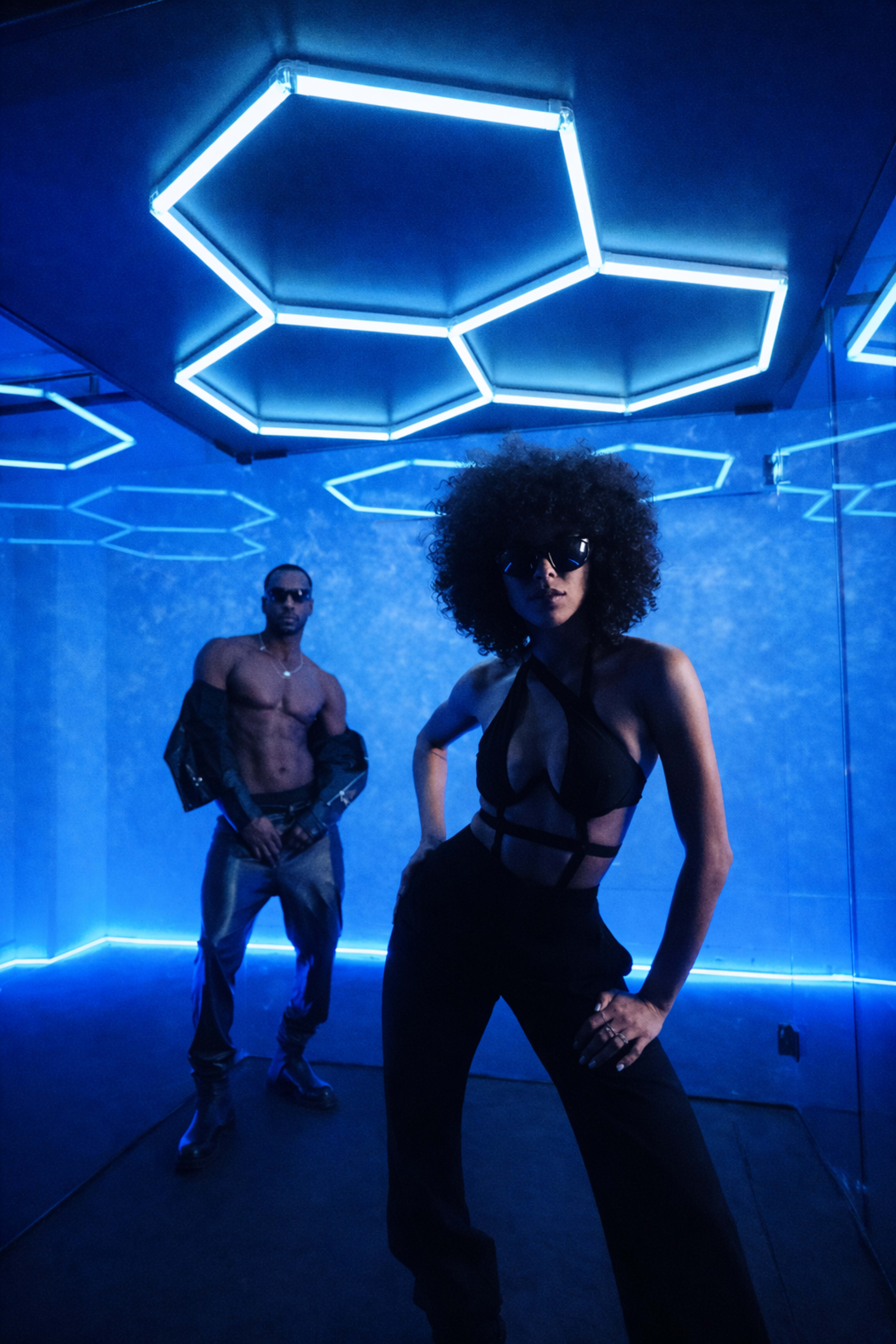 A man stands behind a woman in a stylish outfit, both posed in a room illuminated by blue neon hexagonal lights.