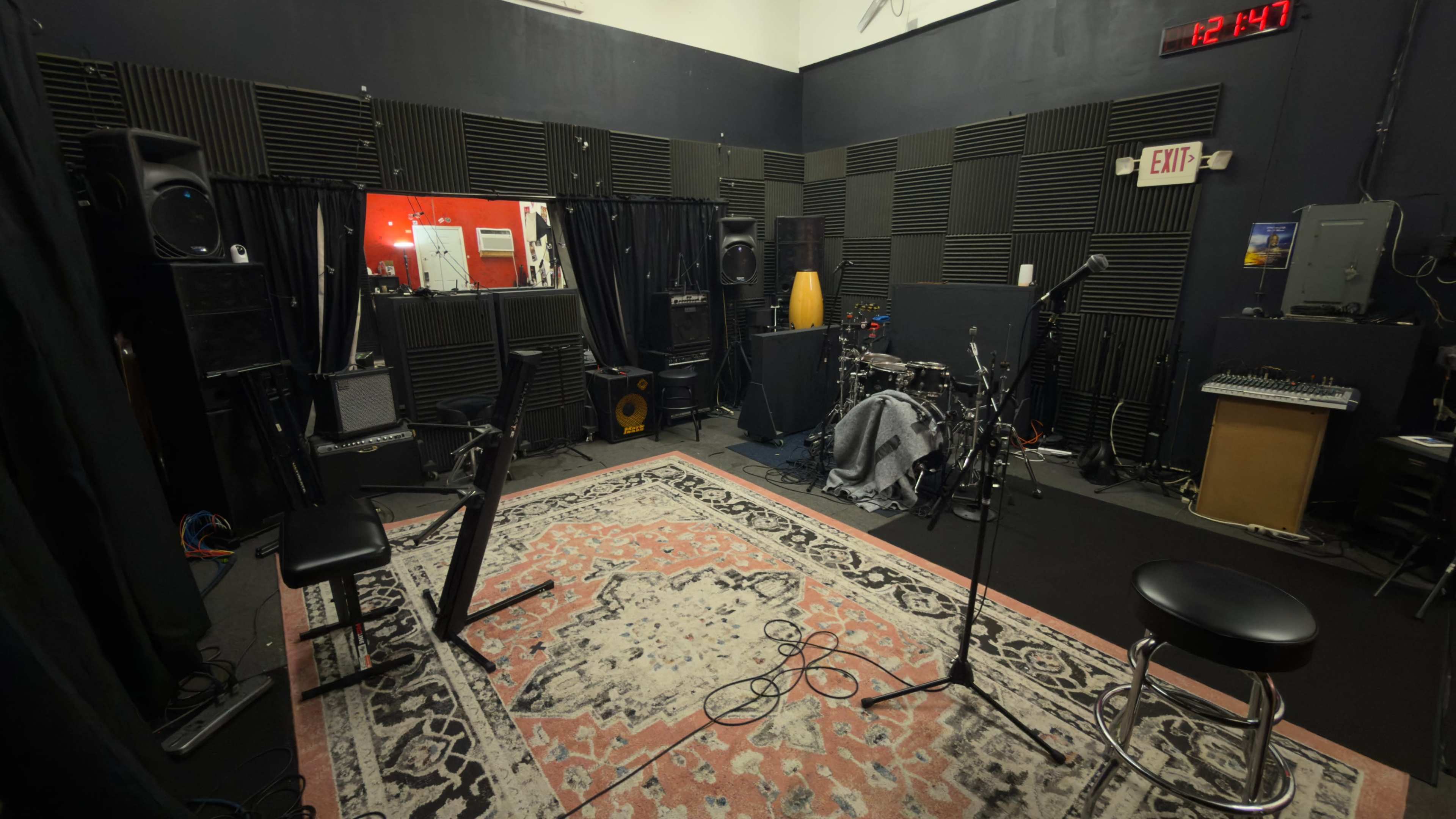The image shows a rehearsal studio equipped with a drum set, amplifiers, and a mixing console, featuring black soundproofing panels and a patterned rug on the floor.