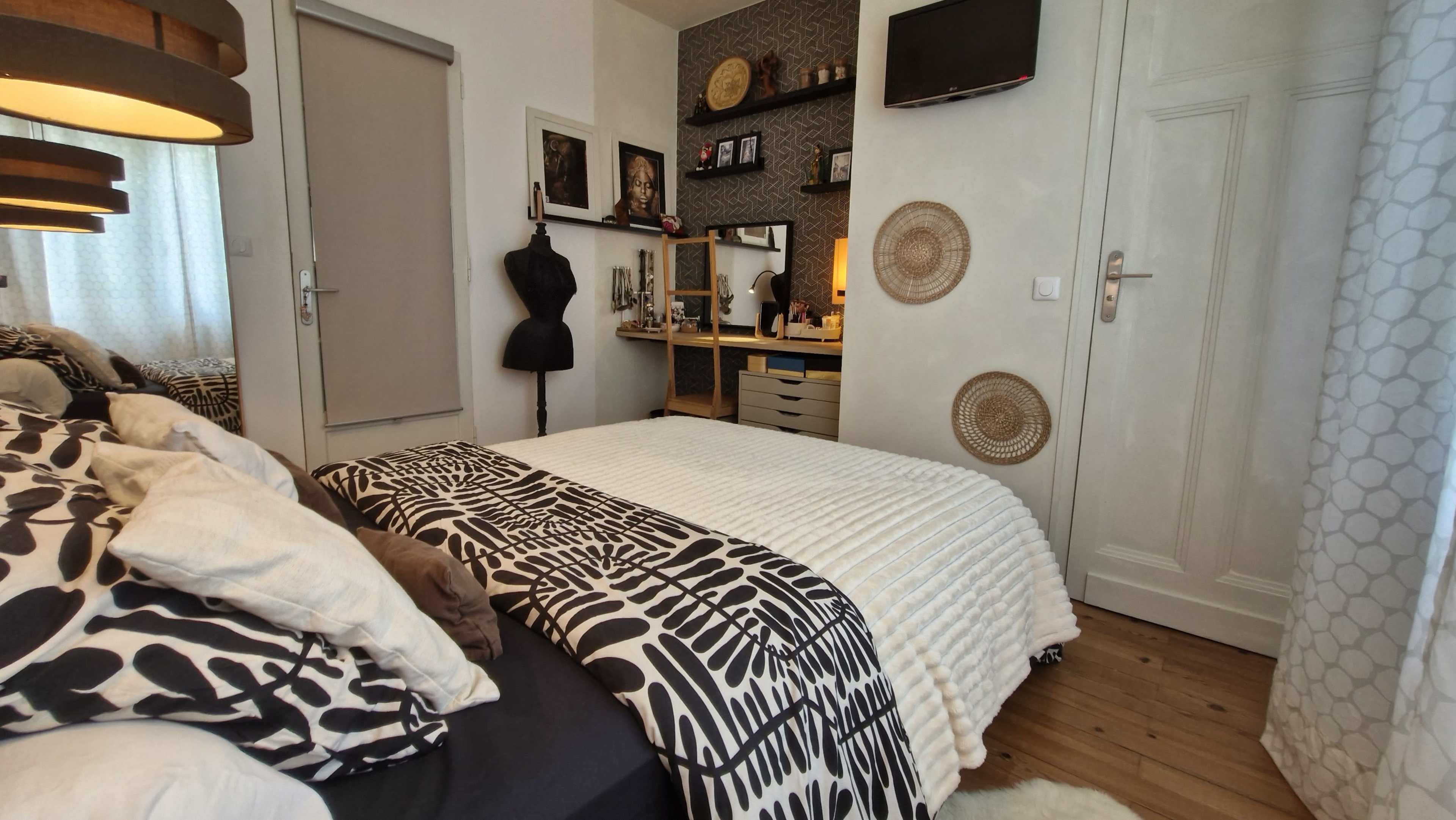 The image shows a stylish bedroom featuring a neatly made bed with patterned bedding, a workspace with a mirror and shelves on one side, and a variety of decorative elements on the walls.