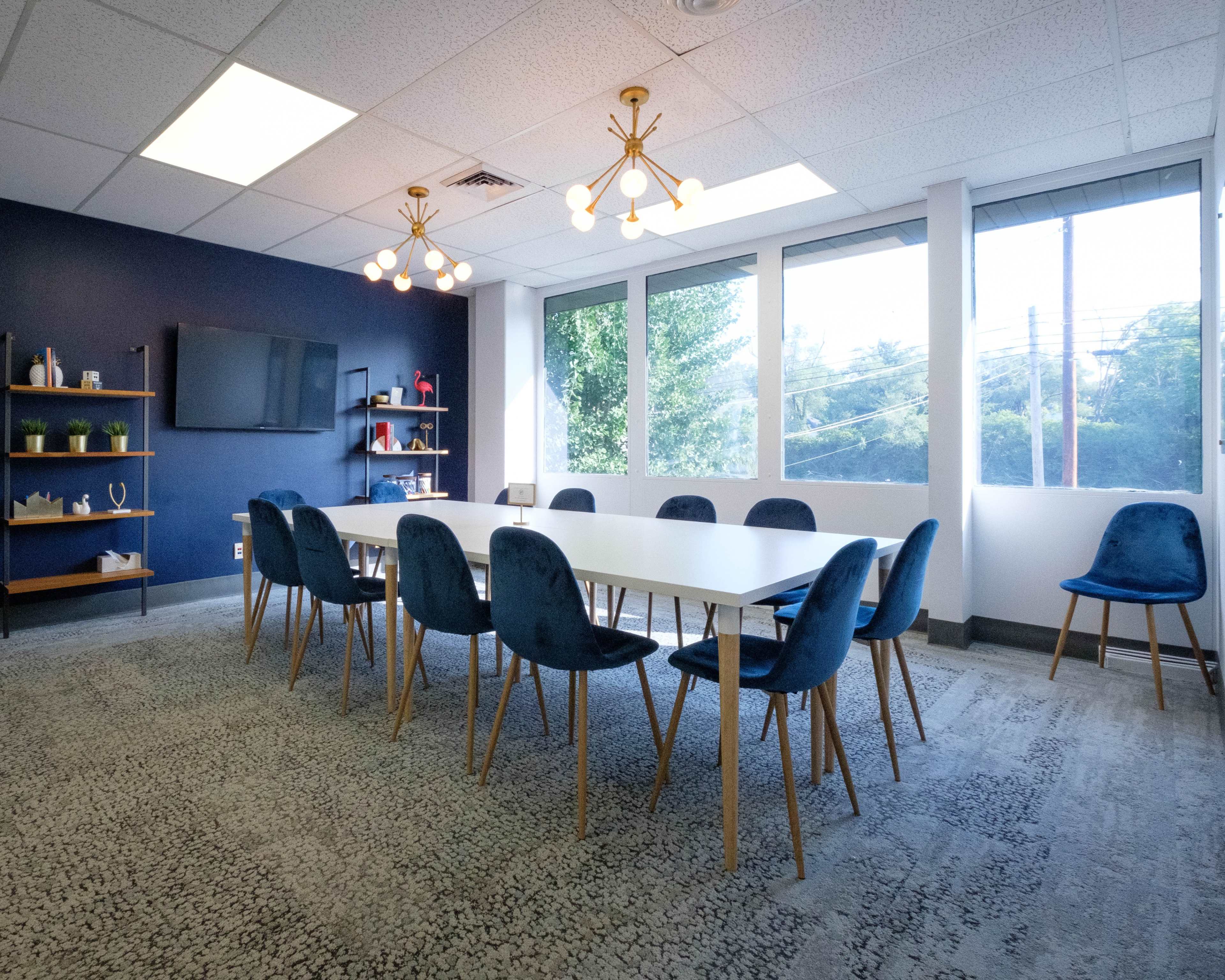 A modern conference room featuring a long white table surrounded by blue chairs, with large windows allowing natural light and a television mounted on the wall.