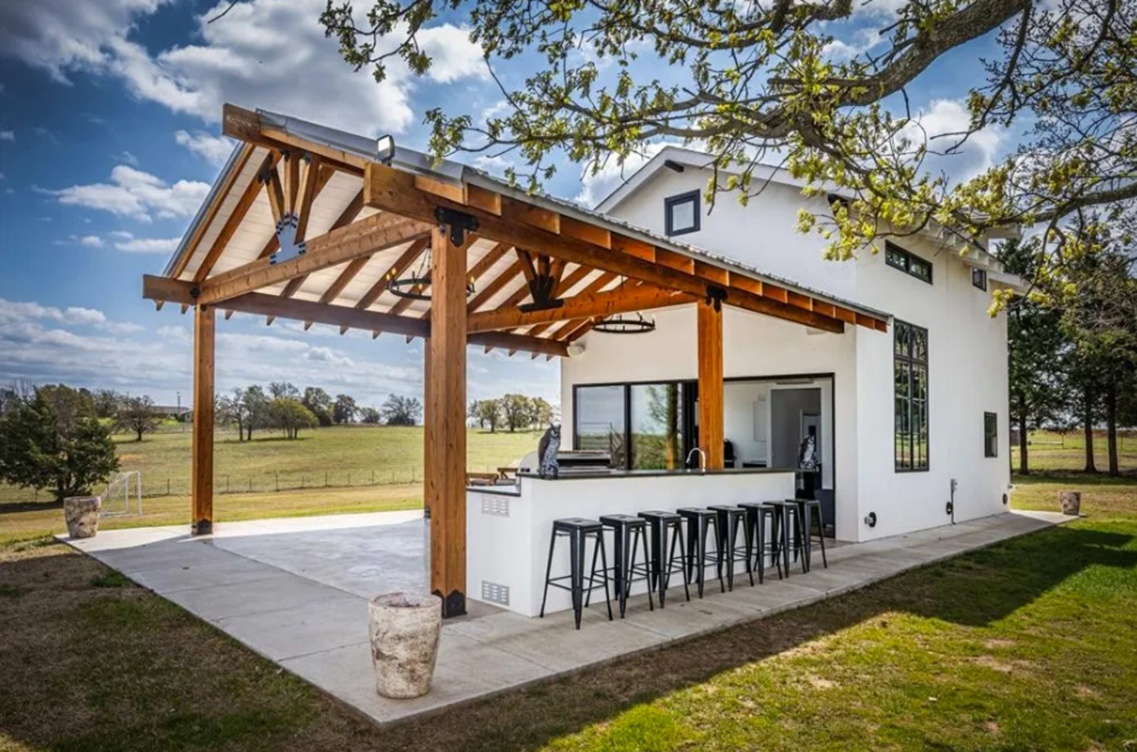 The image shows a modern white house with a large wooden overhang, featuring a bar area and several black stools on a concrete patio, set in a grassy landscape with a view of rolling fields.