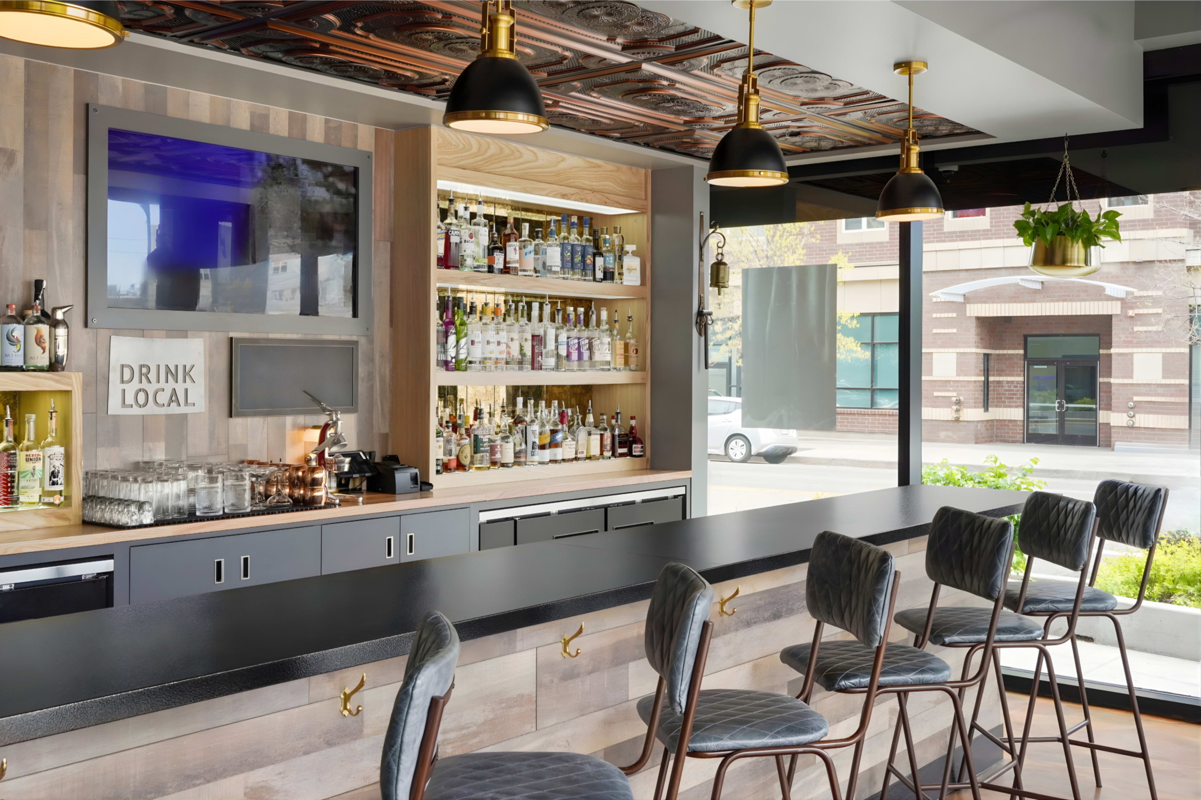 A modern bar features a long counter with high stools, a display of various liquor bottles on shelves, and a television mounted on the wall.