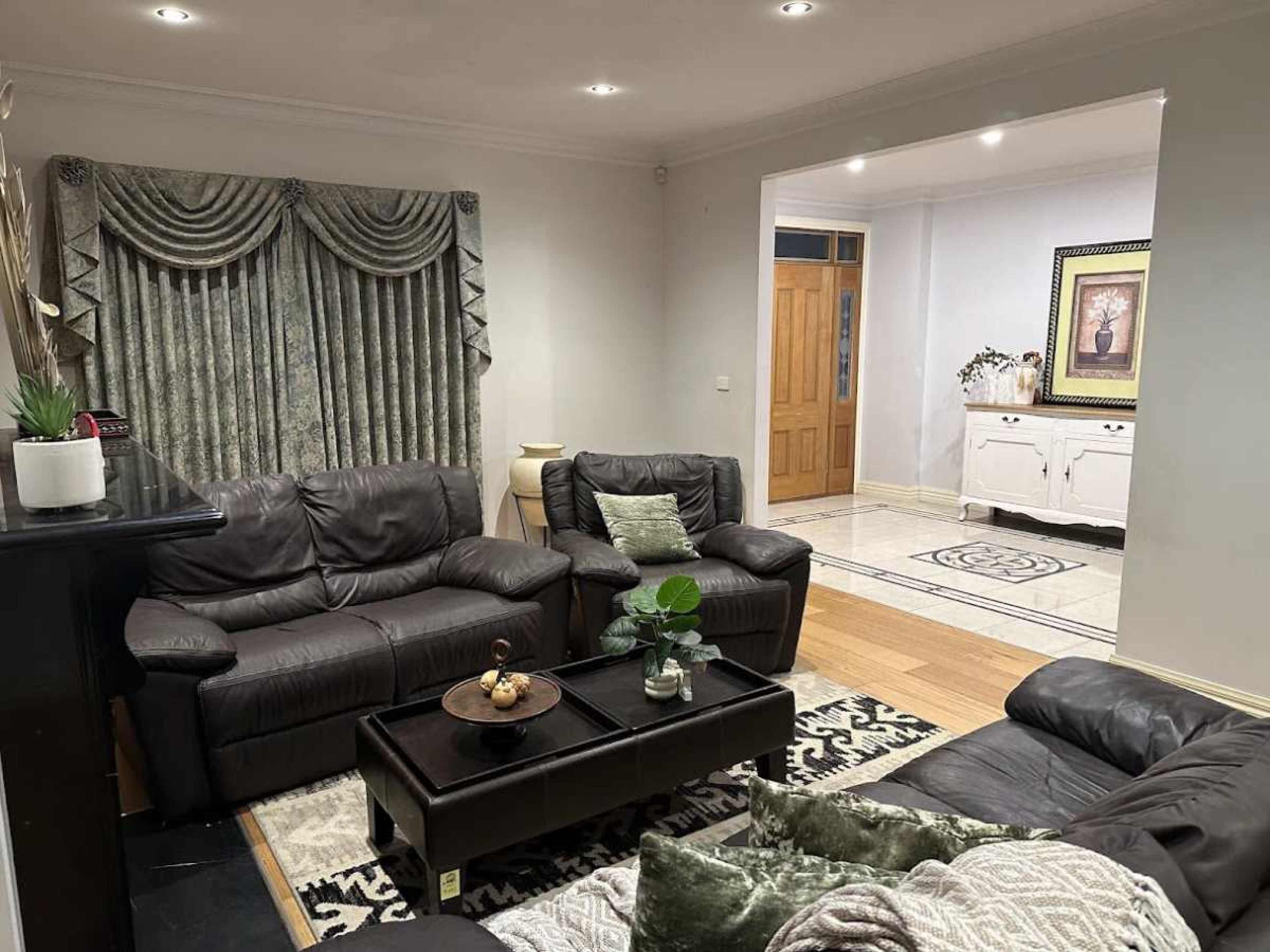 The image shows a living room with dark leather sofas, a coffee table, and a decorative curtain, leading to a hallway with light wooden flooring and a door.