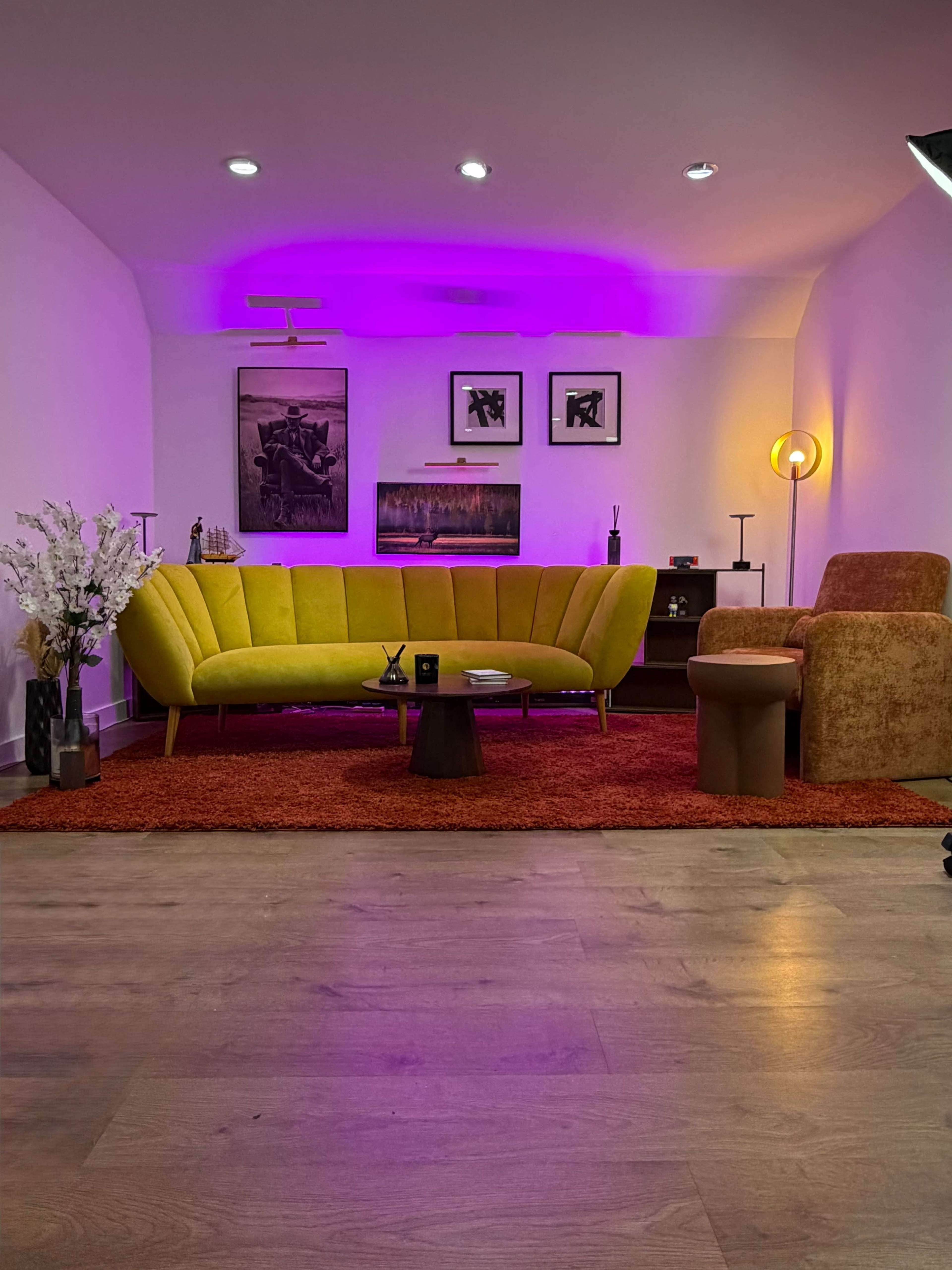 The image shows a modern living room with a yellow curved sofa, a small round table, and an orange rug, illuminated by purple lighting and featuring framed artwork on the walls.