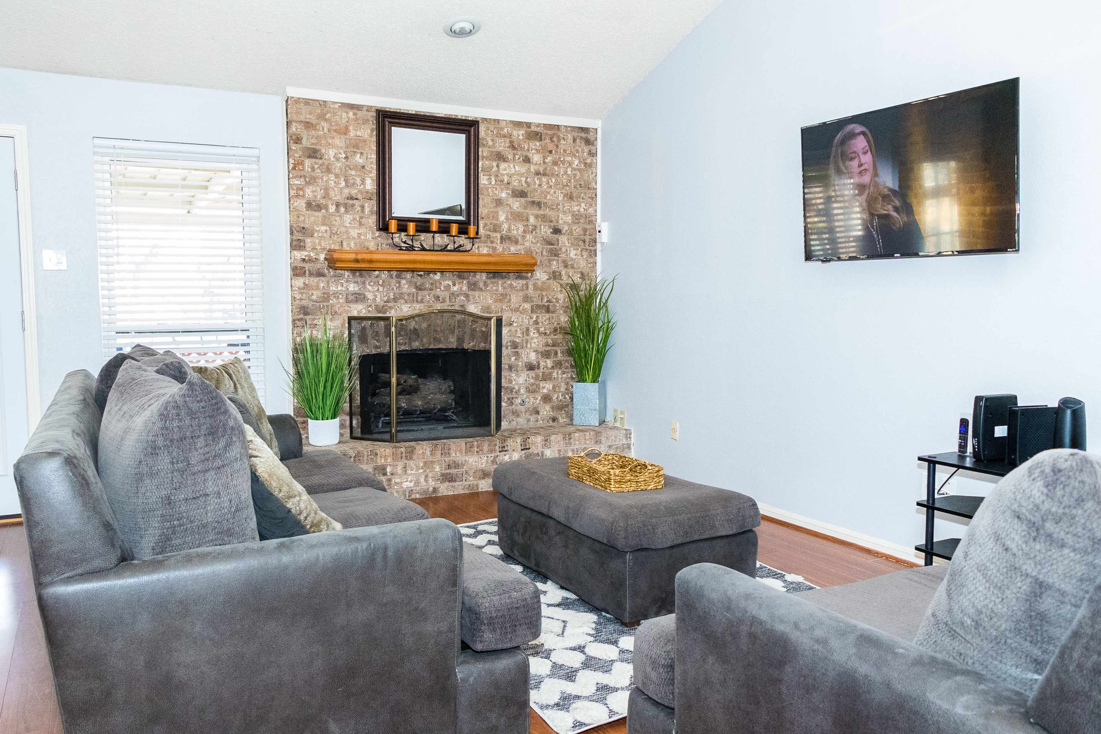 A living room features a brick fireplace, a large gray sectional sofa, and a television mounted on the wall.