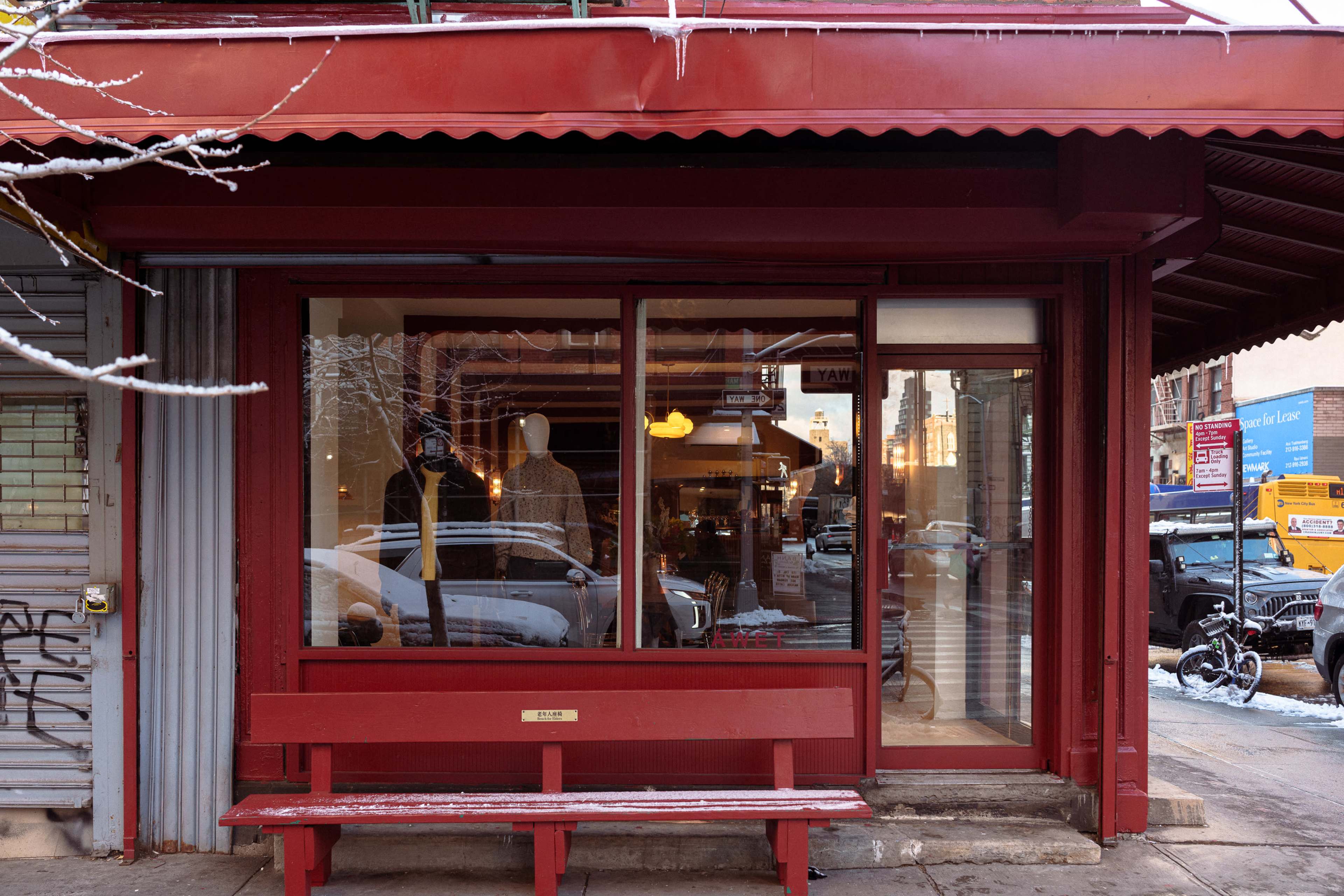 The image shows a red storefront with large windows and a bench outside, located on a city street lined with parked cars and pedestrians.