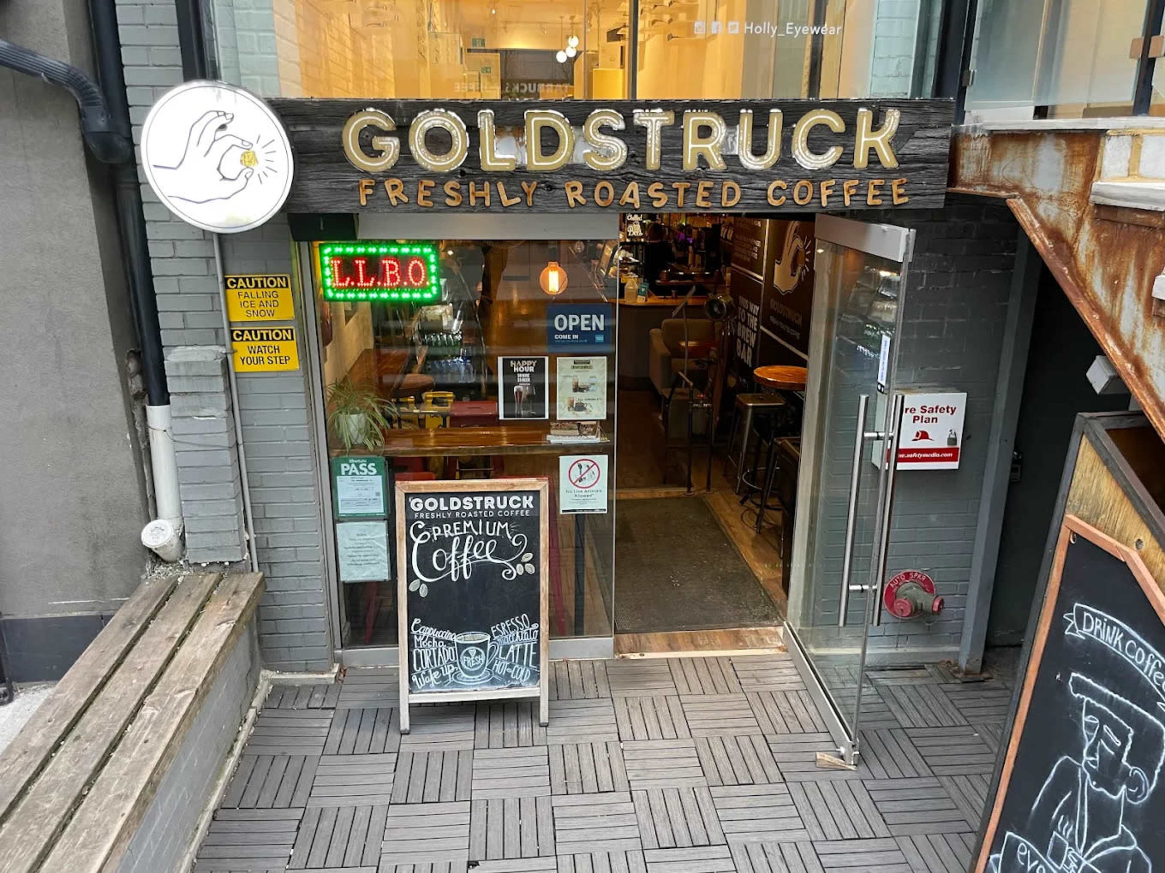 The entrance of a coffee shop named "Goldstruck" features an open sign, a menu board, and warning signs near the door.