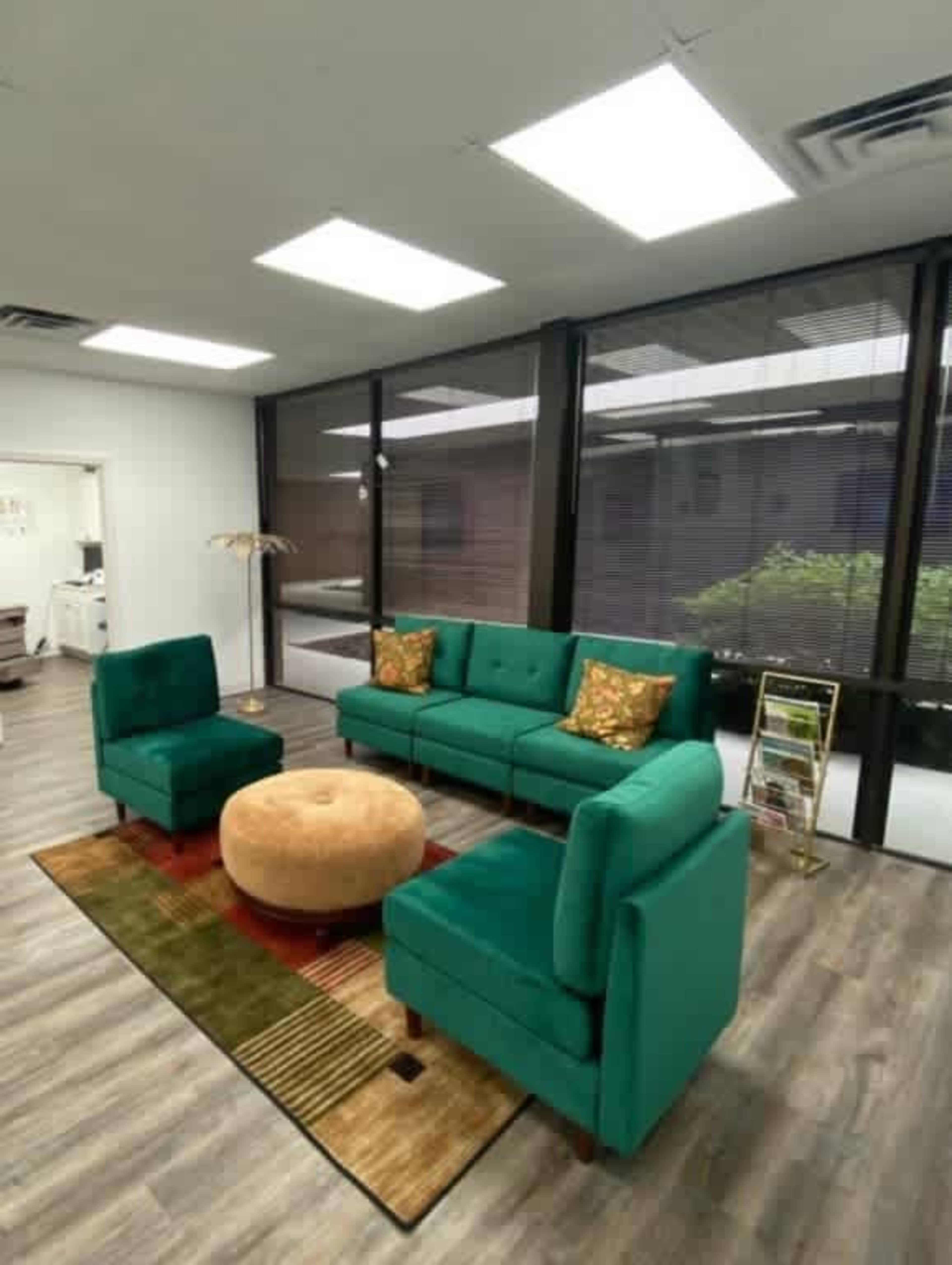 The image shows a modern sitting area featuring a green sofa with decorative pillows, two matching chairs, a round ottoman, and large windows letting in natural light.