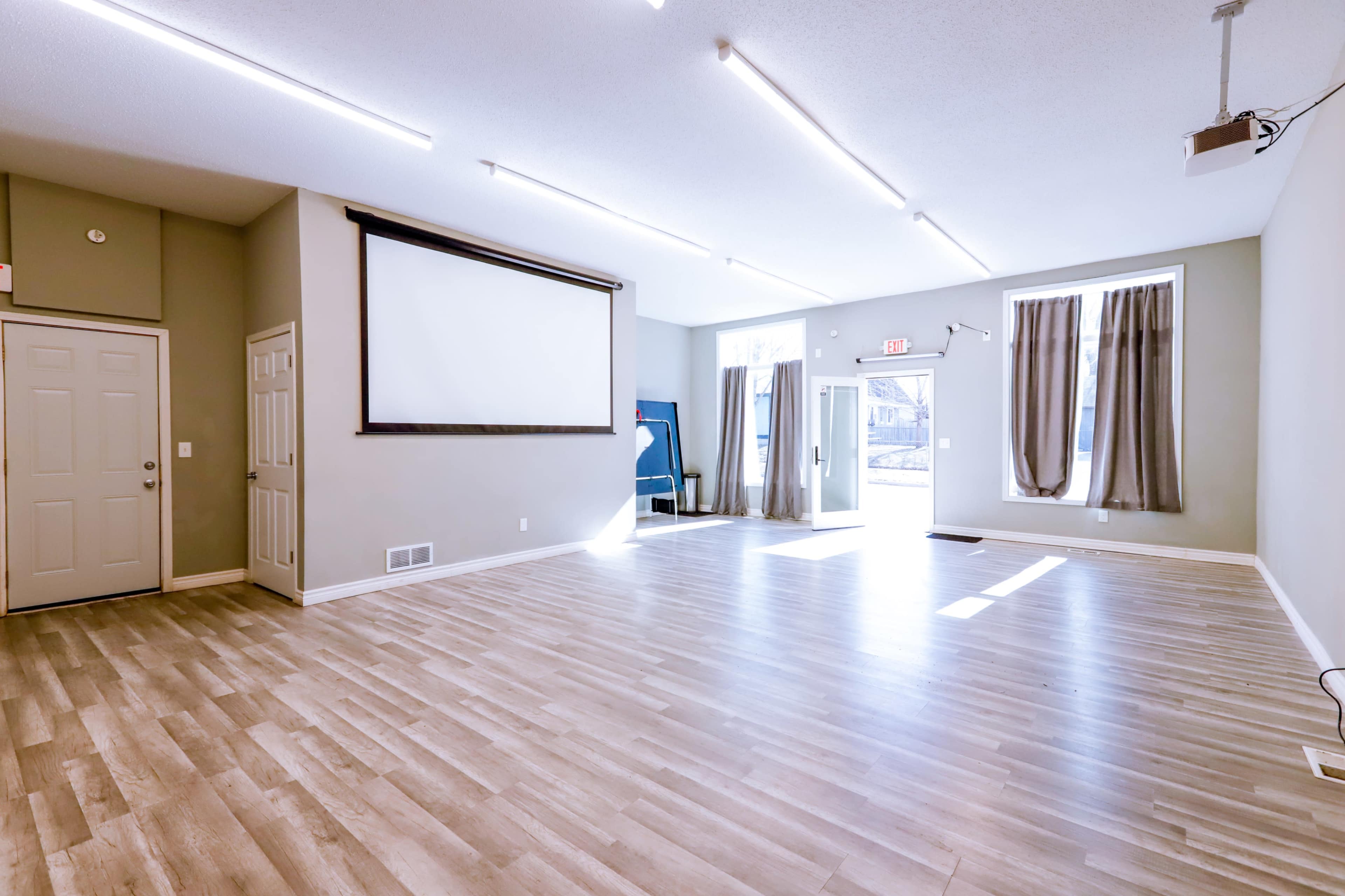 A spacious, well-lit room with wooden flooring, a projector screen mounted on the wall, and large windows with curtains.