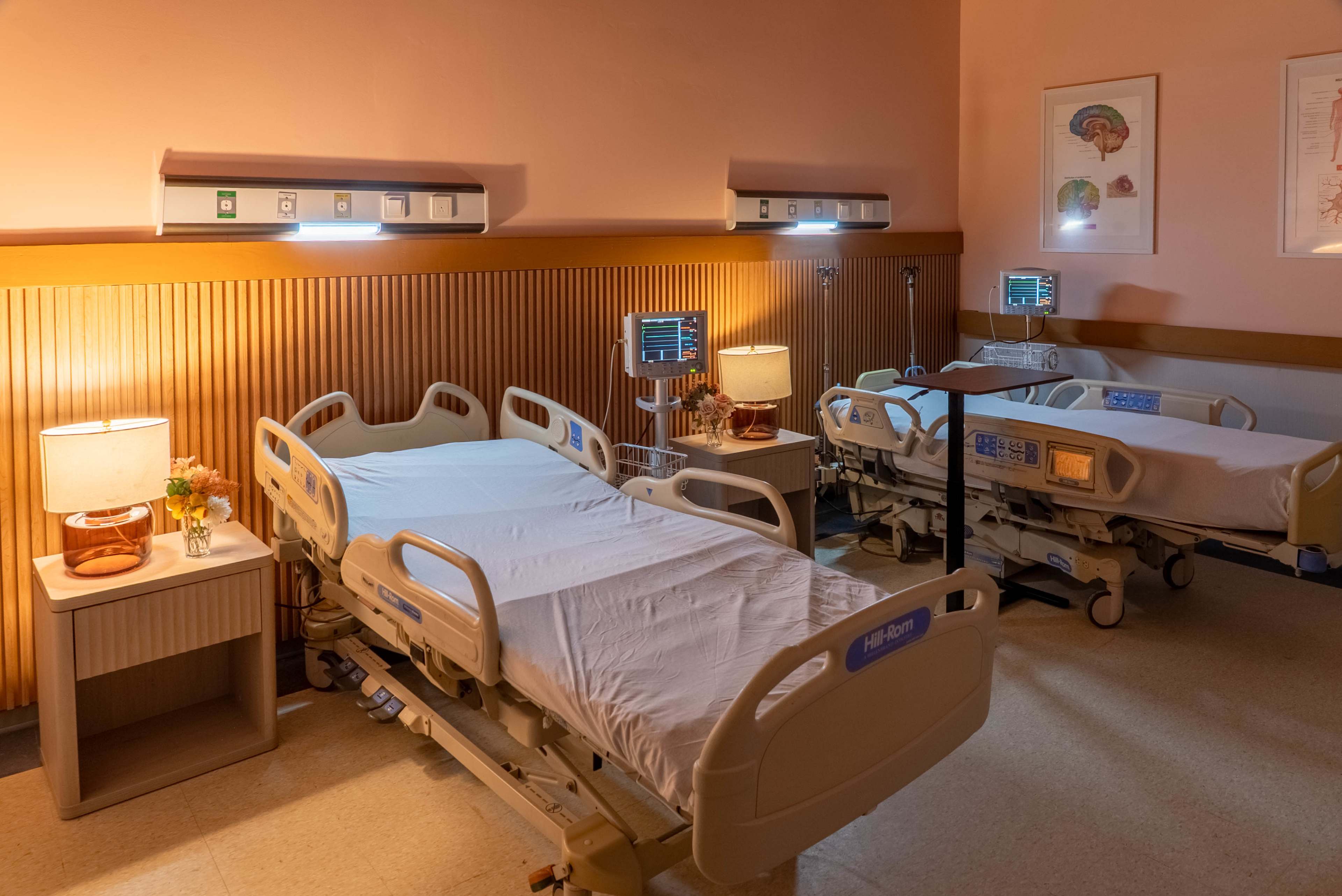 The image shows a hospital room with three adjustable beds, bedside tables with lamps, and medical monitors on the walls.