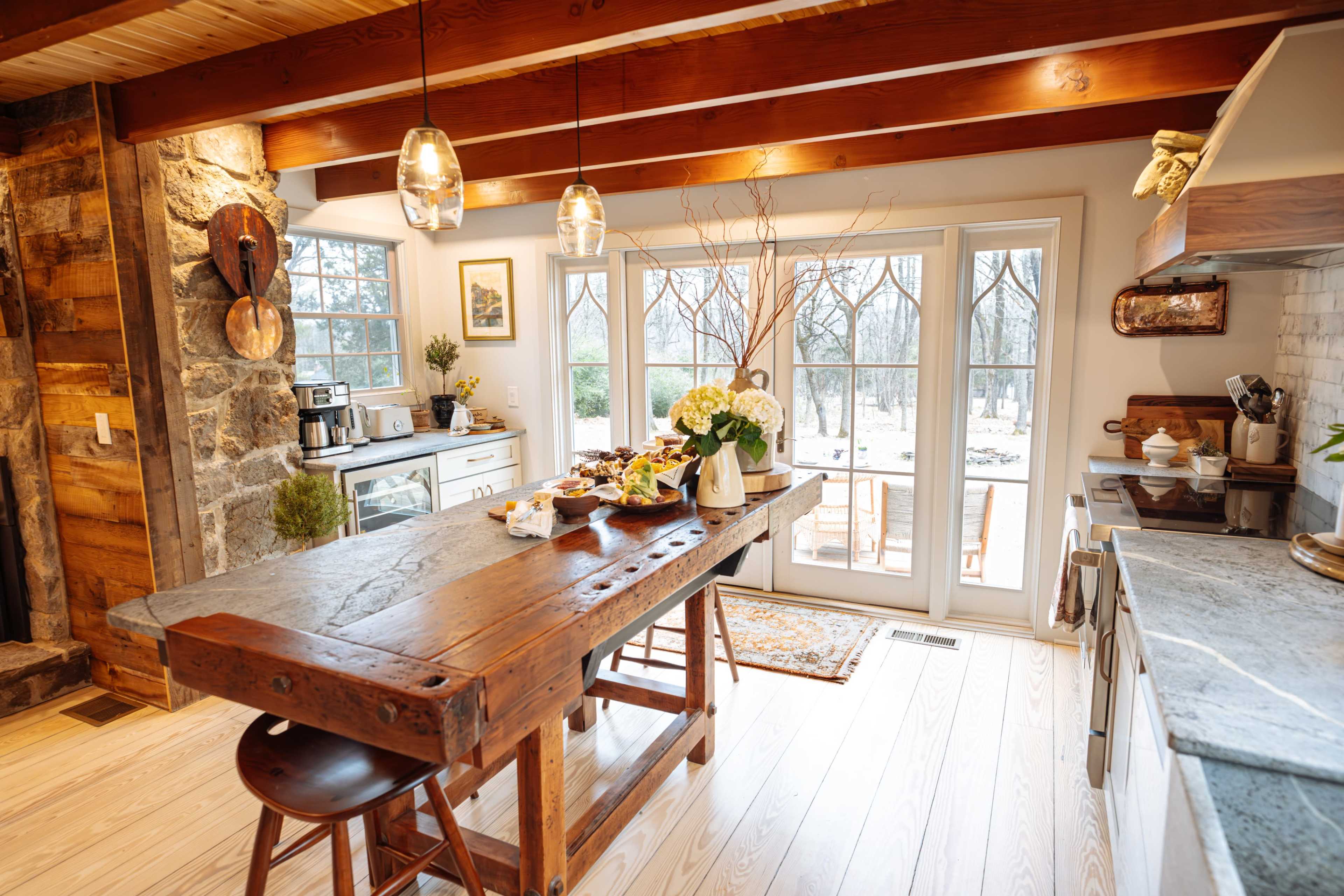 A rustic kitchen features a large wooden table set with food, surrounded by windows that provide a view of an outdoor landscape.