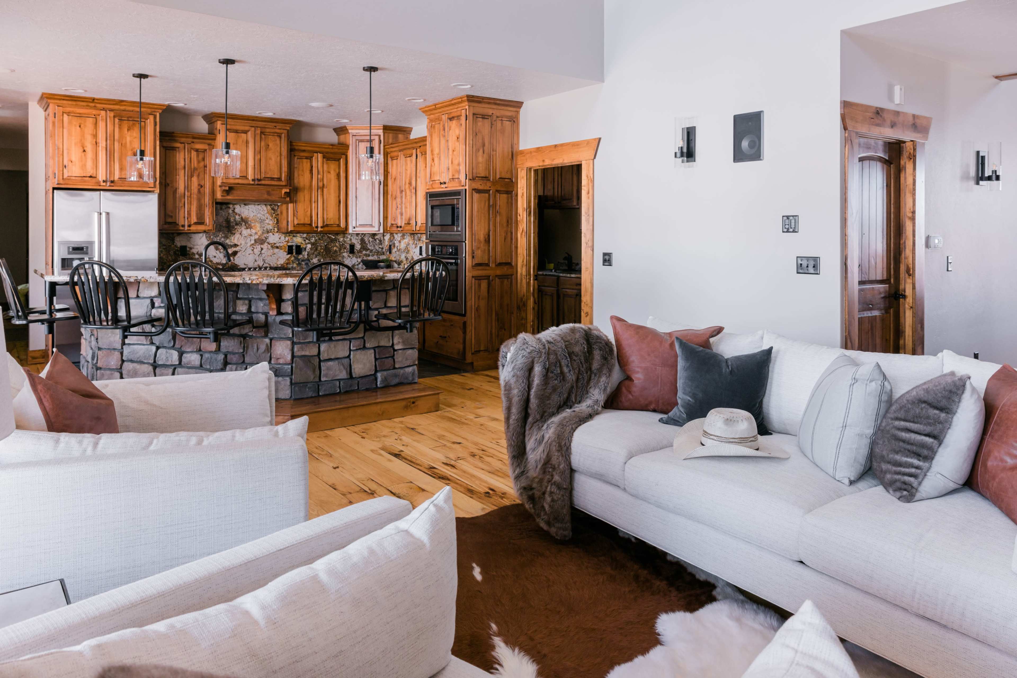 A modern living room with a light-colored sectional sofa, a large rug, and a kitchen featuring wooden cabinets and a stone bar.