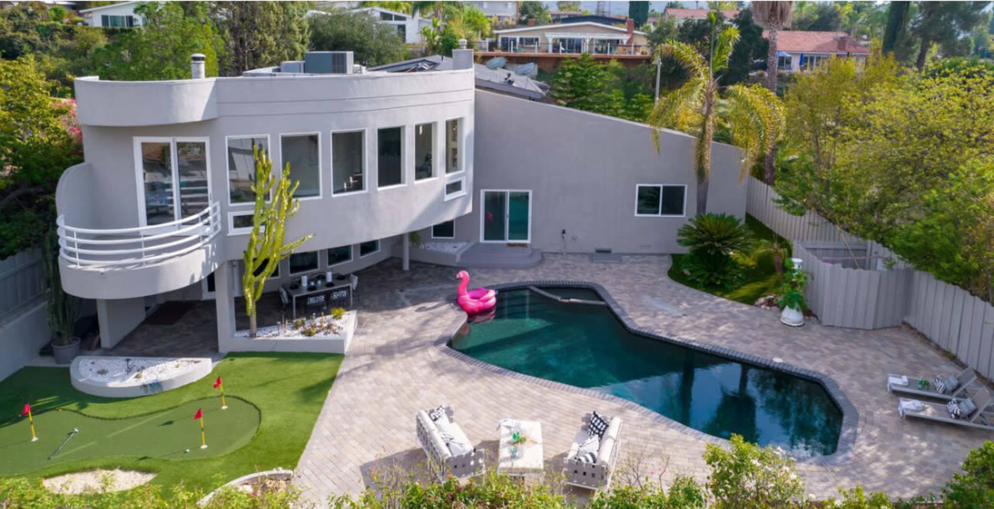 A modern house with a pool, a golf putting green, and a pink flamingo float in the backyard, surrounded by greenery.