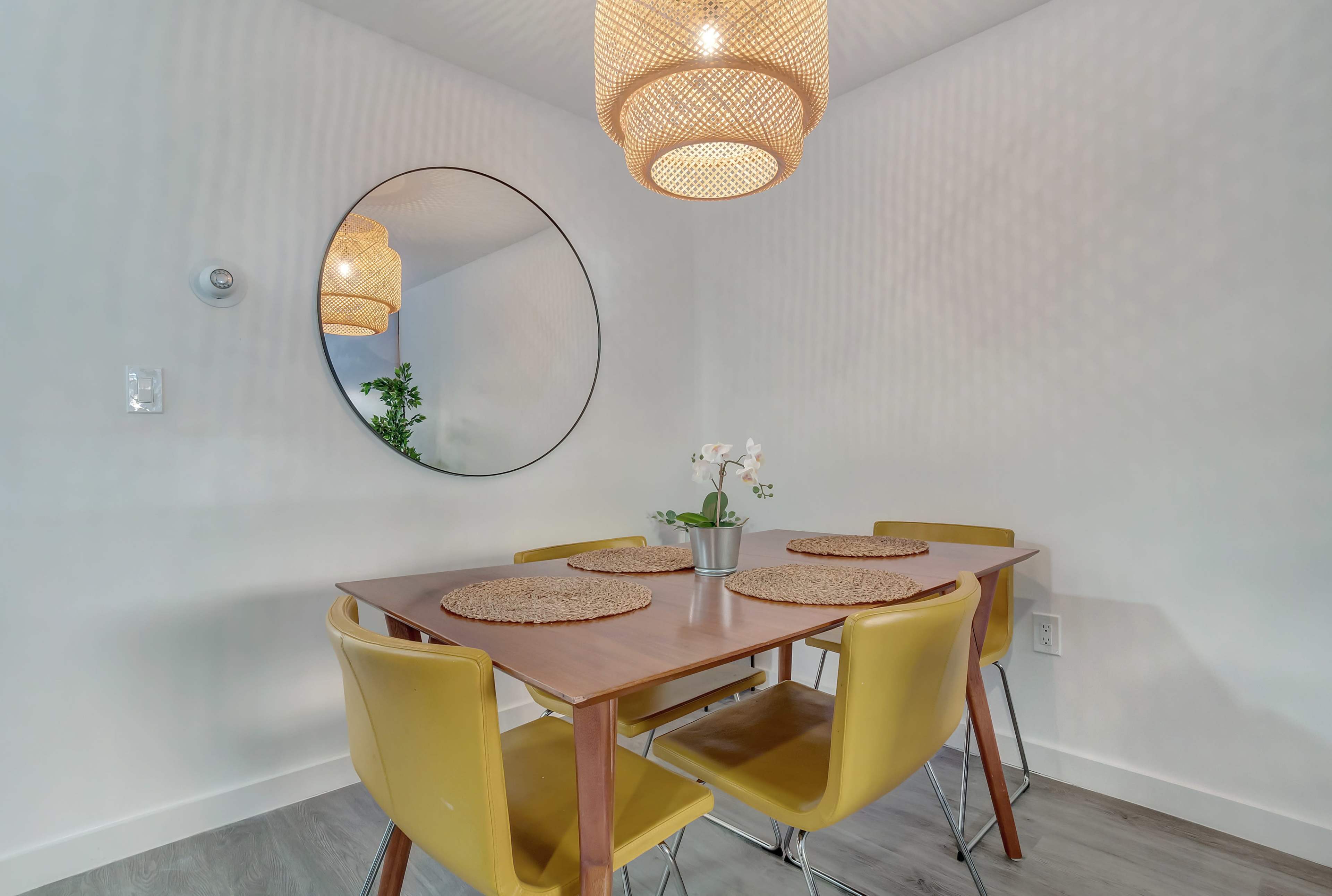 A dining area features a wooden table with yellow chairs, a round mirror on the wall, and a small potted plant at the center.