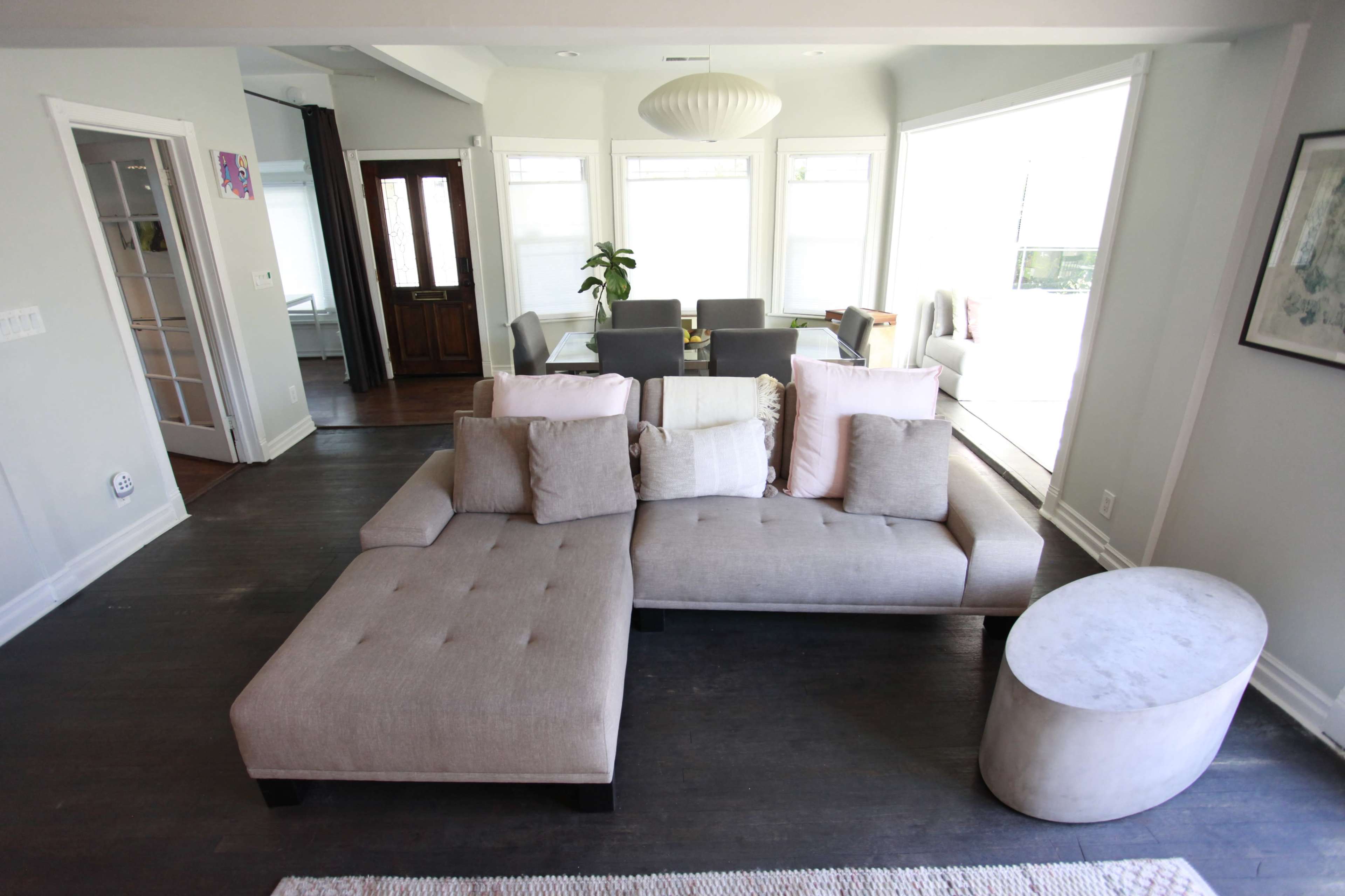 A modern living room features a beige sectional sofa facing a dining area with a table and chairs, surrounded by large windows.