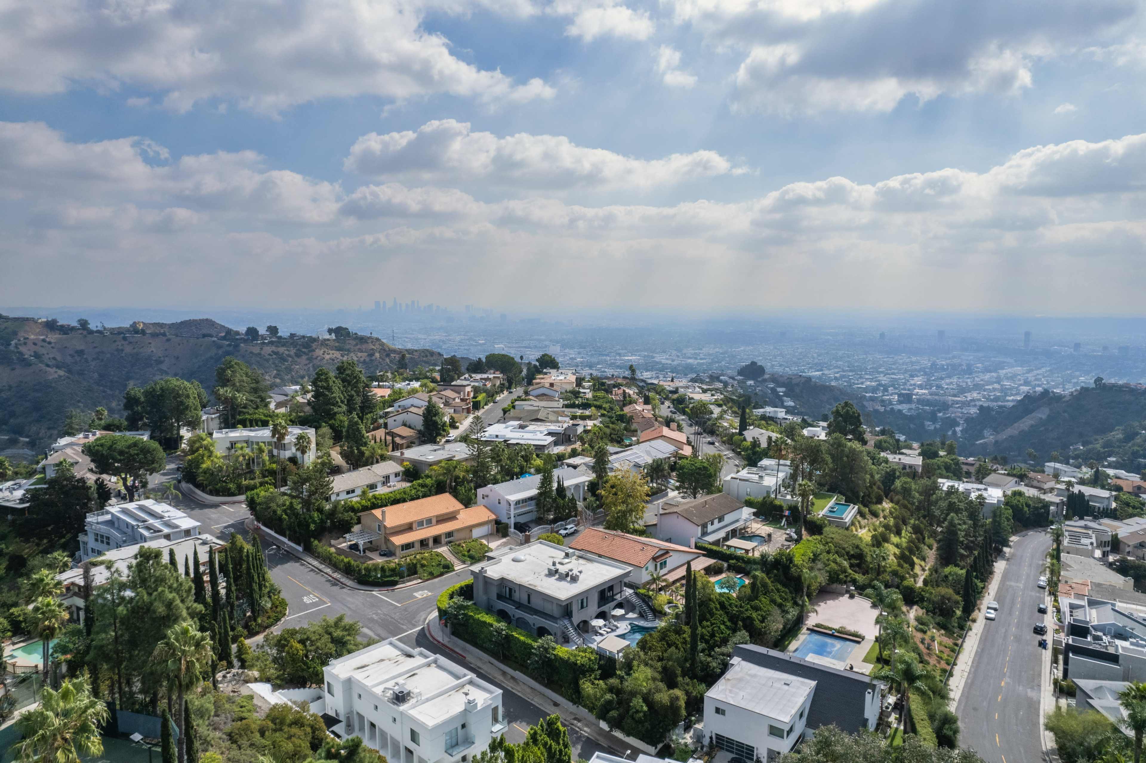 Aerial view of a residential area in a hilly landscape, featuring various houses with pools and greenery, overlooking a city skyline in the distance.