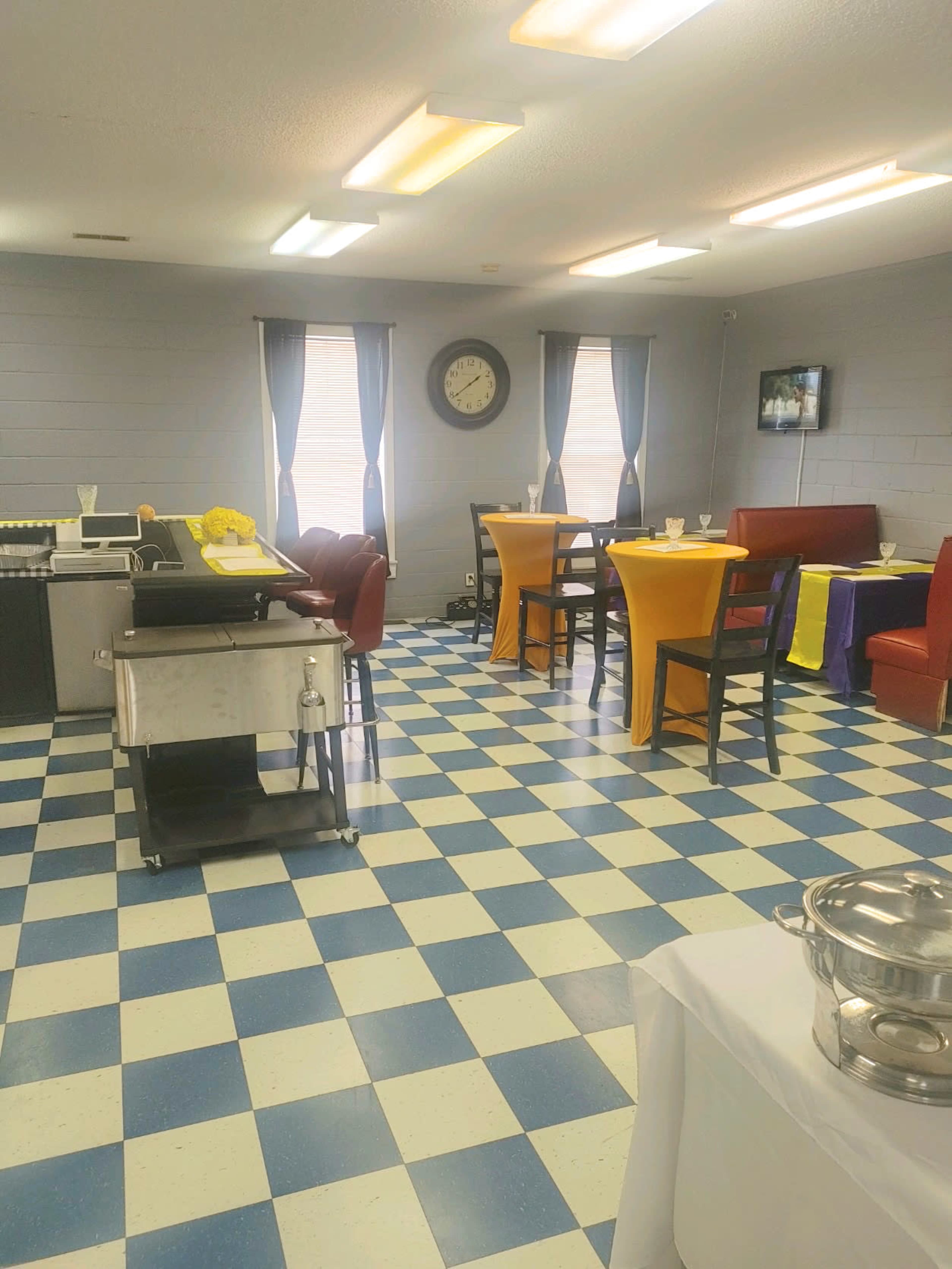 A dining area with a checkered blue and white floor, tables dressed in yellow and purple tablecloths, and a clock on the wall.