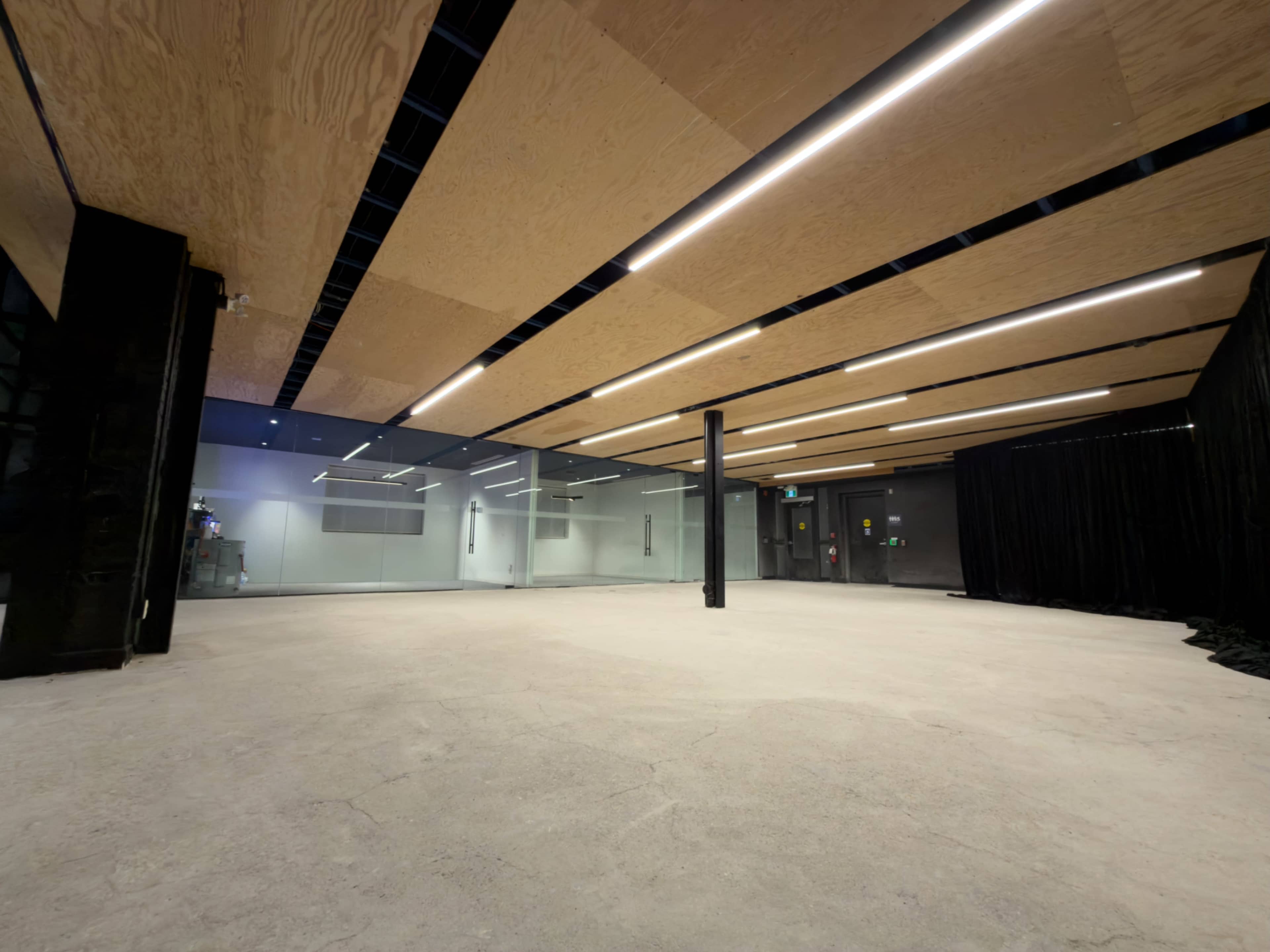 The image shows a spacious, empty room with a concrete floor and wooden ceiling panels, featuring large glass walls on one side.