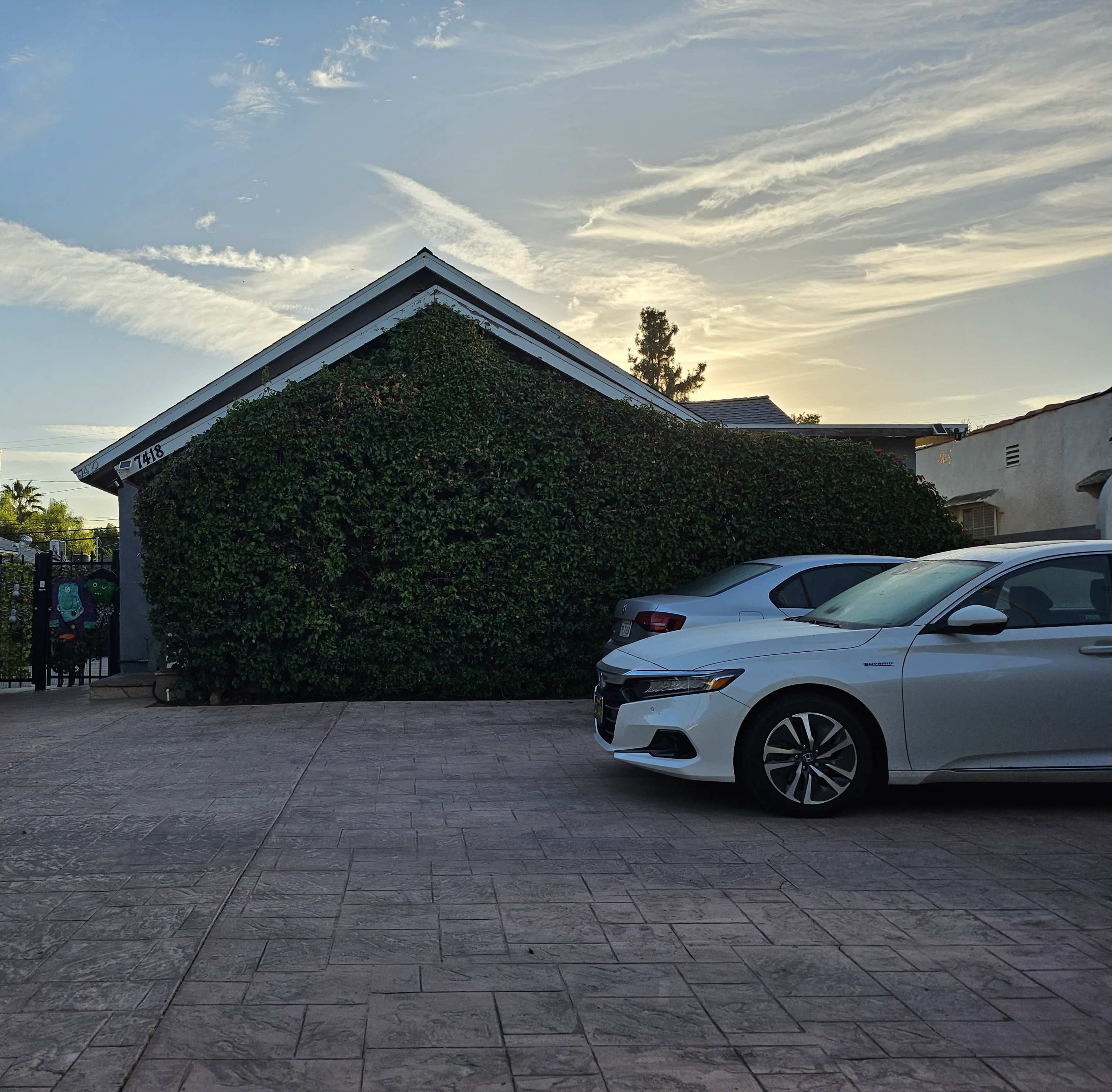 A house covered in greenery stands next to two parked cars in a paved driveway under a cloudy sky at sunset.