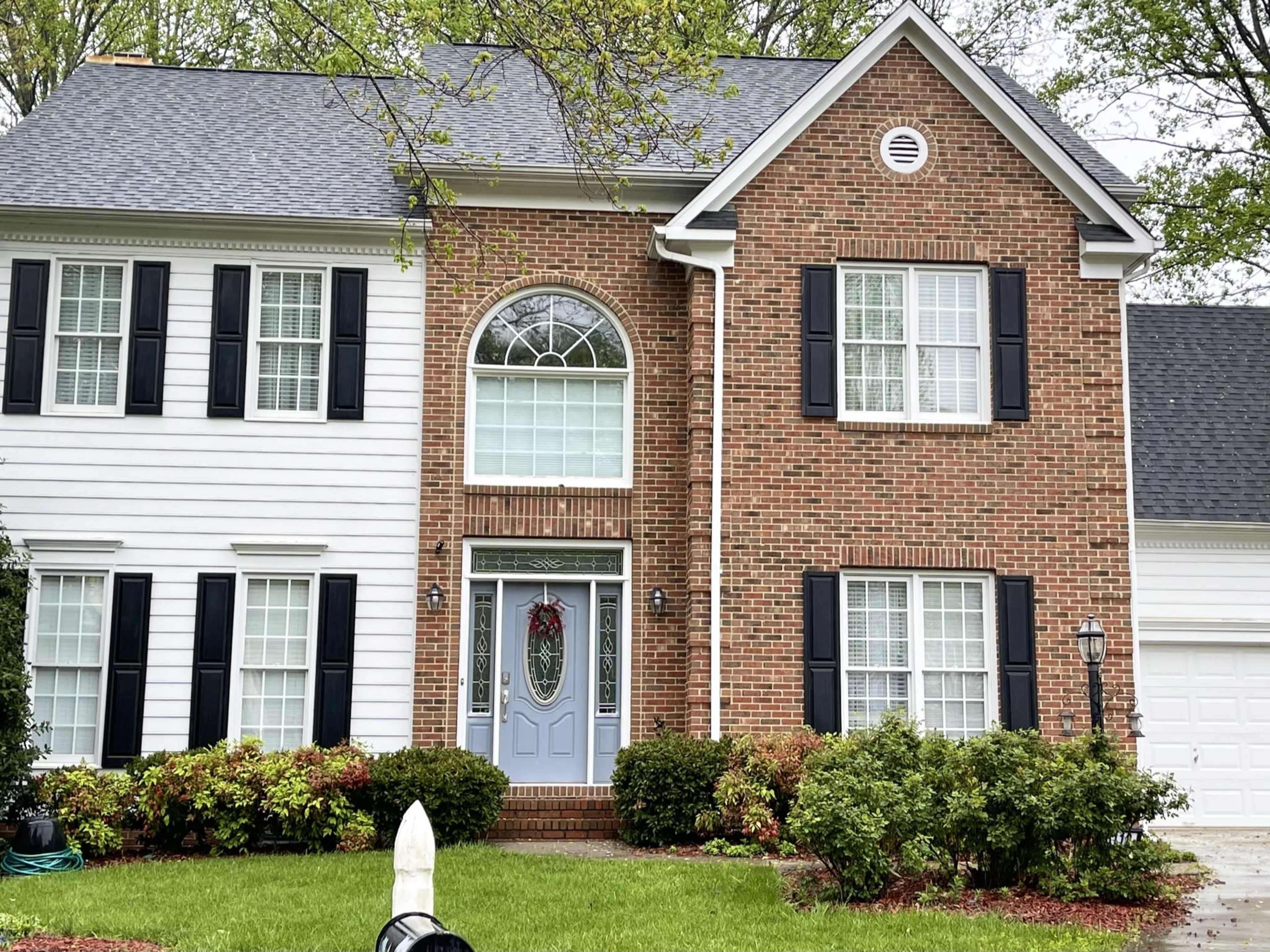 A two-story brick-front house with white siding, black shutters, and a decorative front door surrounded by bushes and a small lawn.