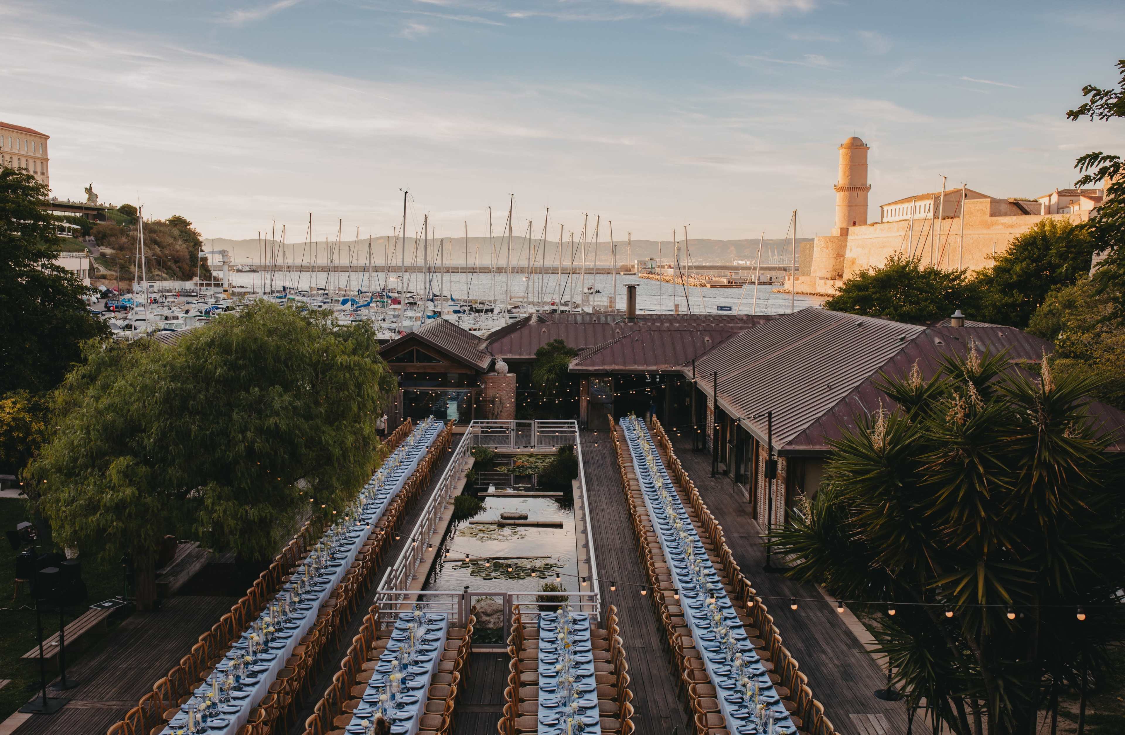 The image shows a long outdoor dining setup beside a marina, with boats in the background and a brick tower visible to the right.