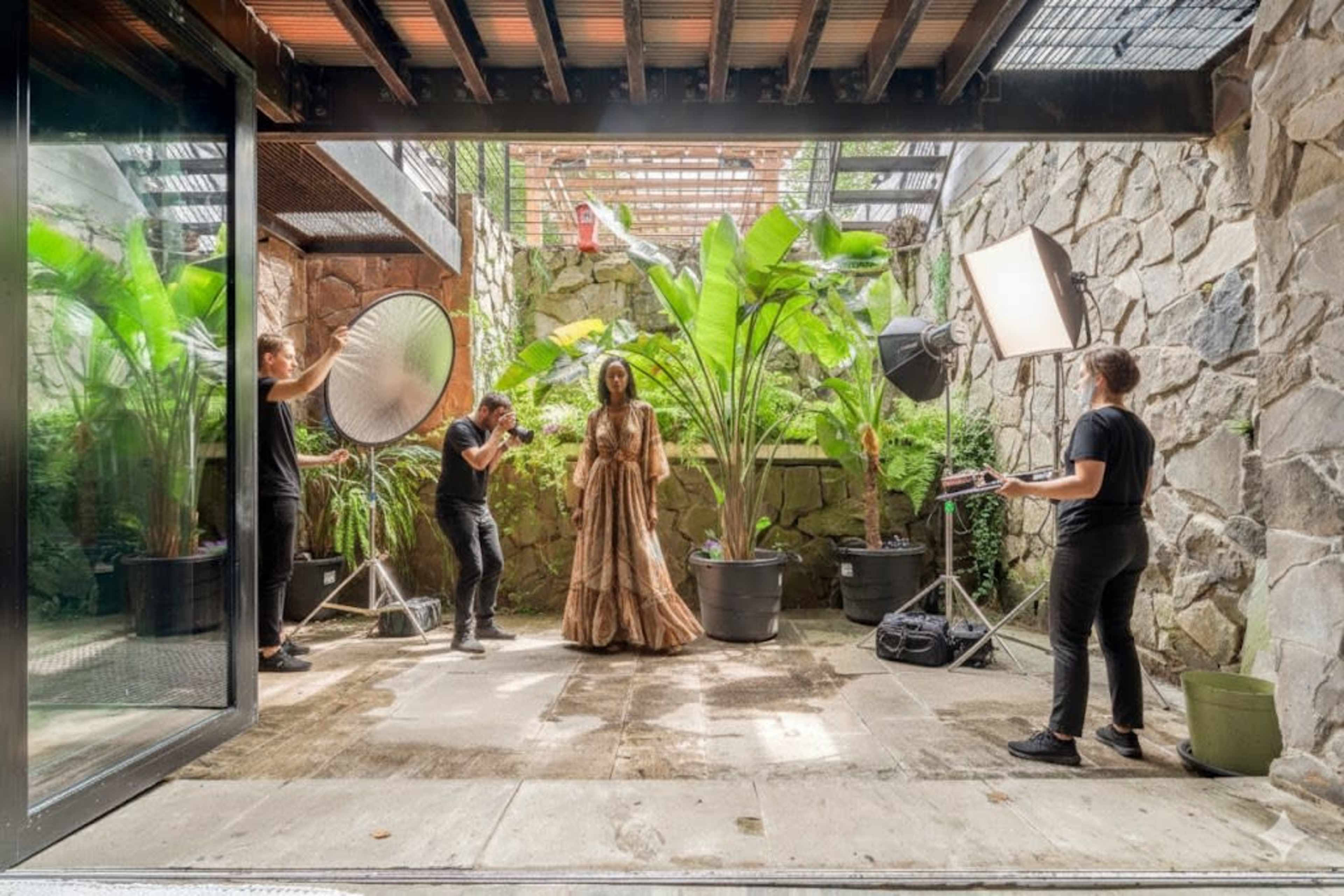 A photoshoot is taking place in an outdoor space surrounded by stone walls and tropical plants, with a model dressed in a long gown and multiple photographers setting up equipment.