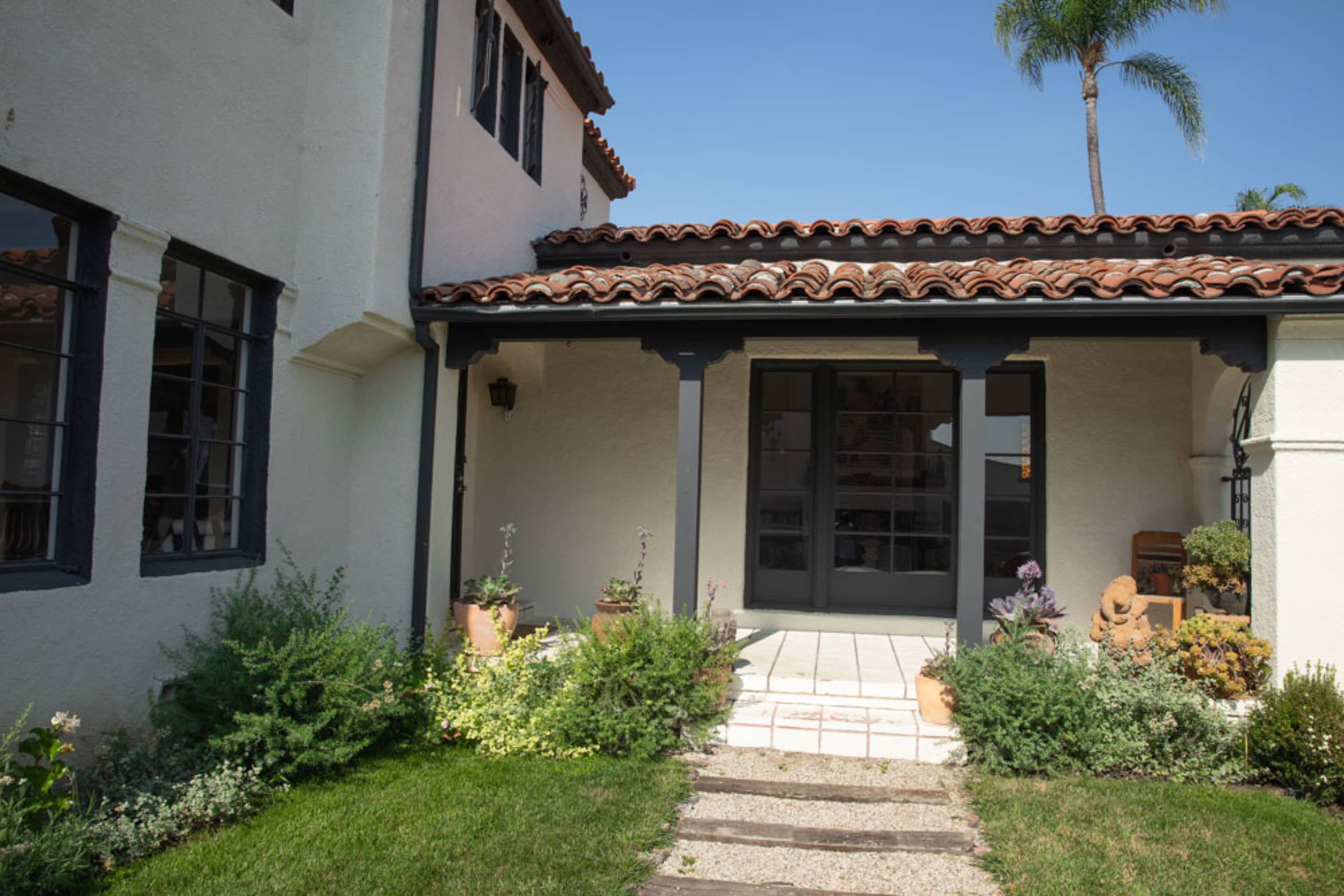 The image shows a well-maintained entryway of a house, featuring a tiled path, potted plants, and a wooden door under a tiled roof.