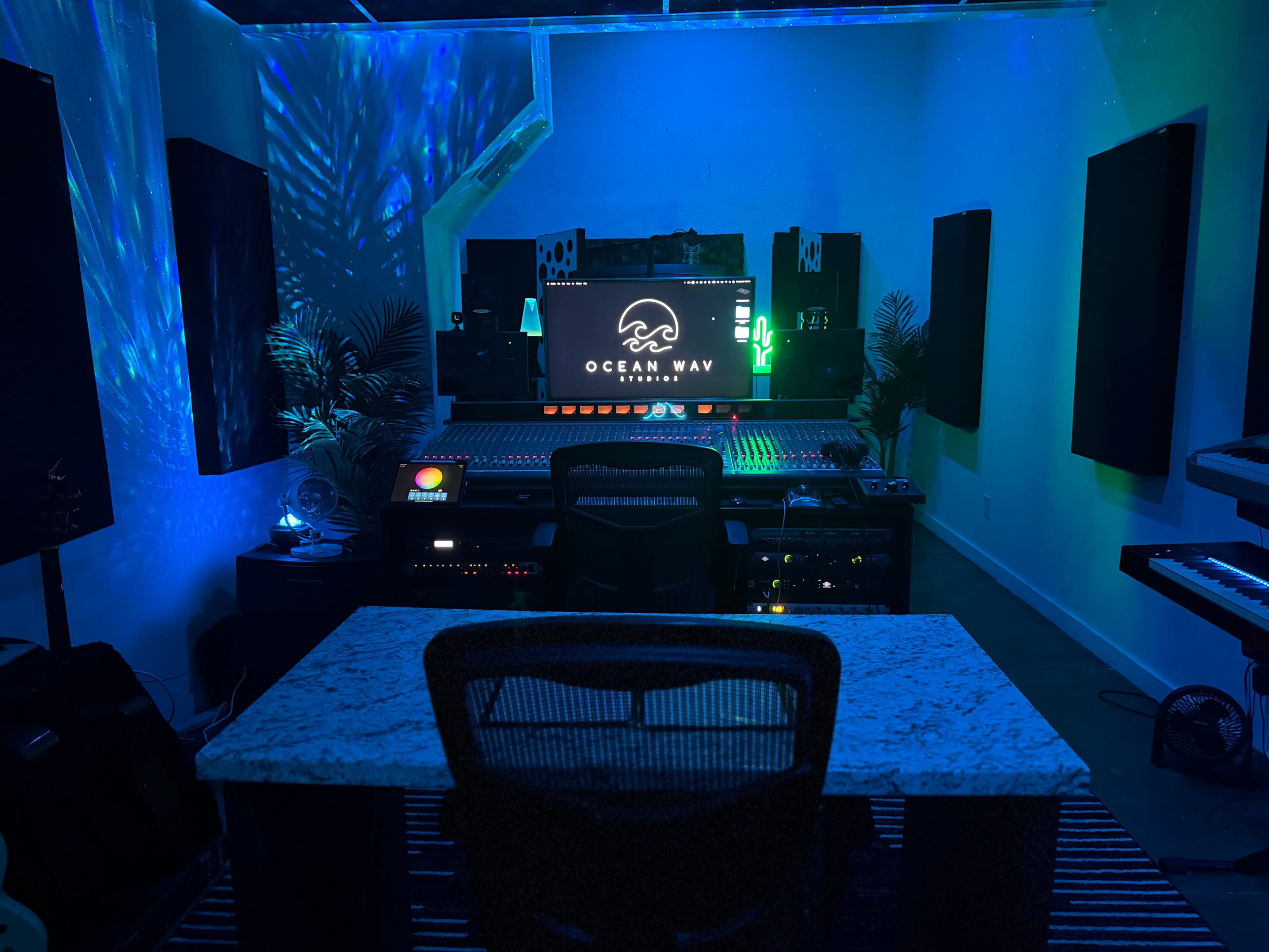 A music studio features a control desk with a computer monitor displaying "OCEAN WAV," surrounded by audio equipment and blue-green ambient lighting.