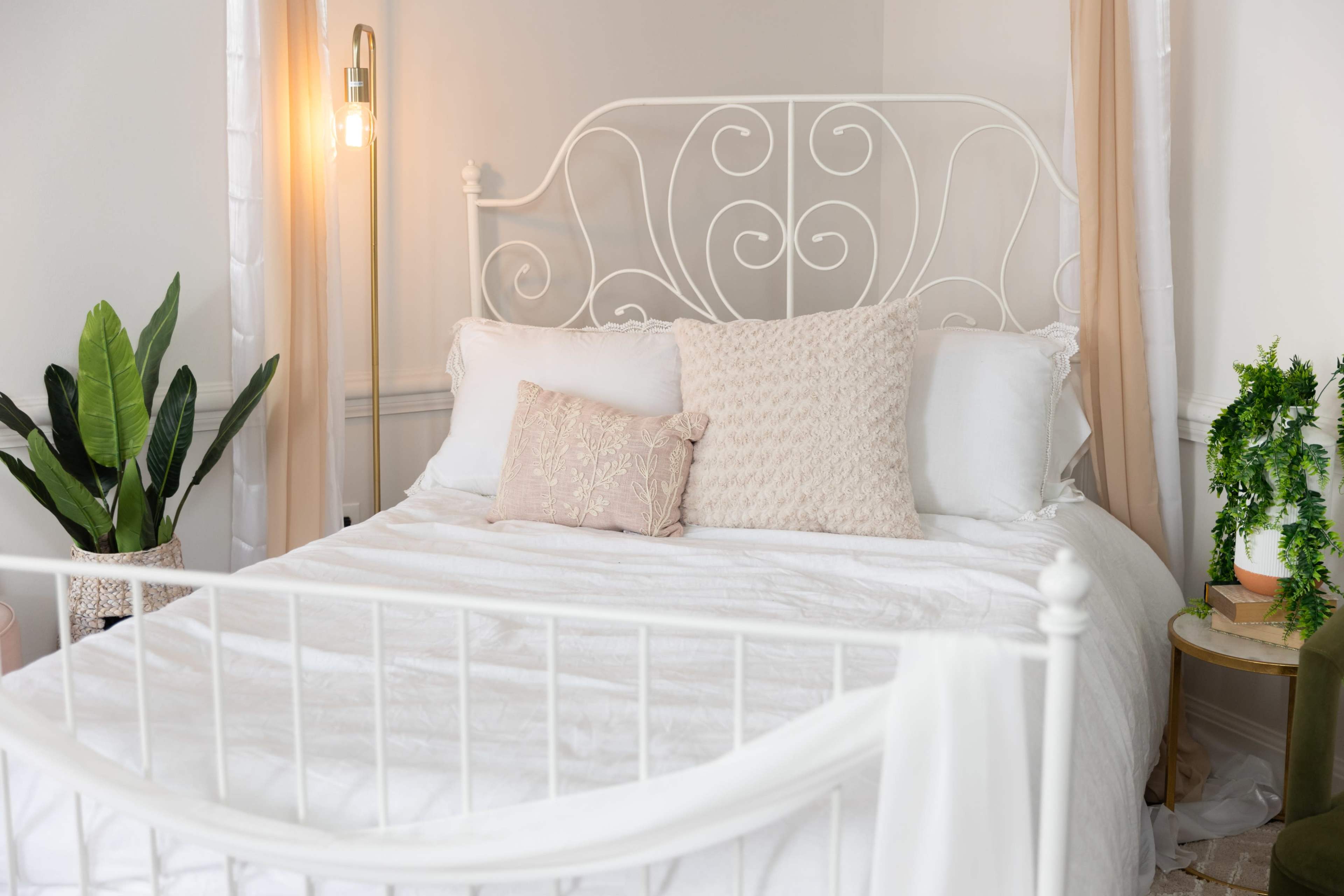 A white metal bed with decorative pillows is set up in a softly lit bedroom featuring indoor plants and a bedside table.
