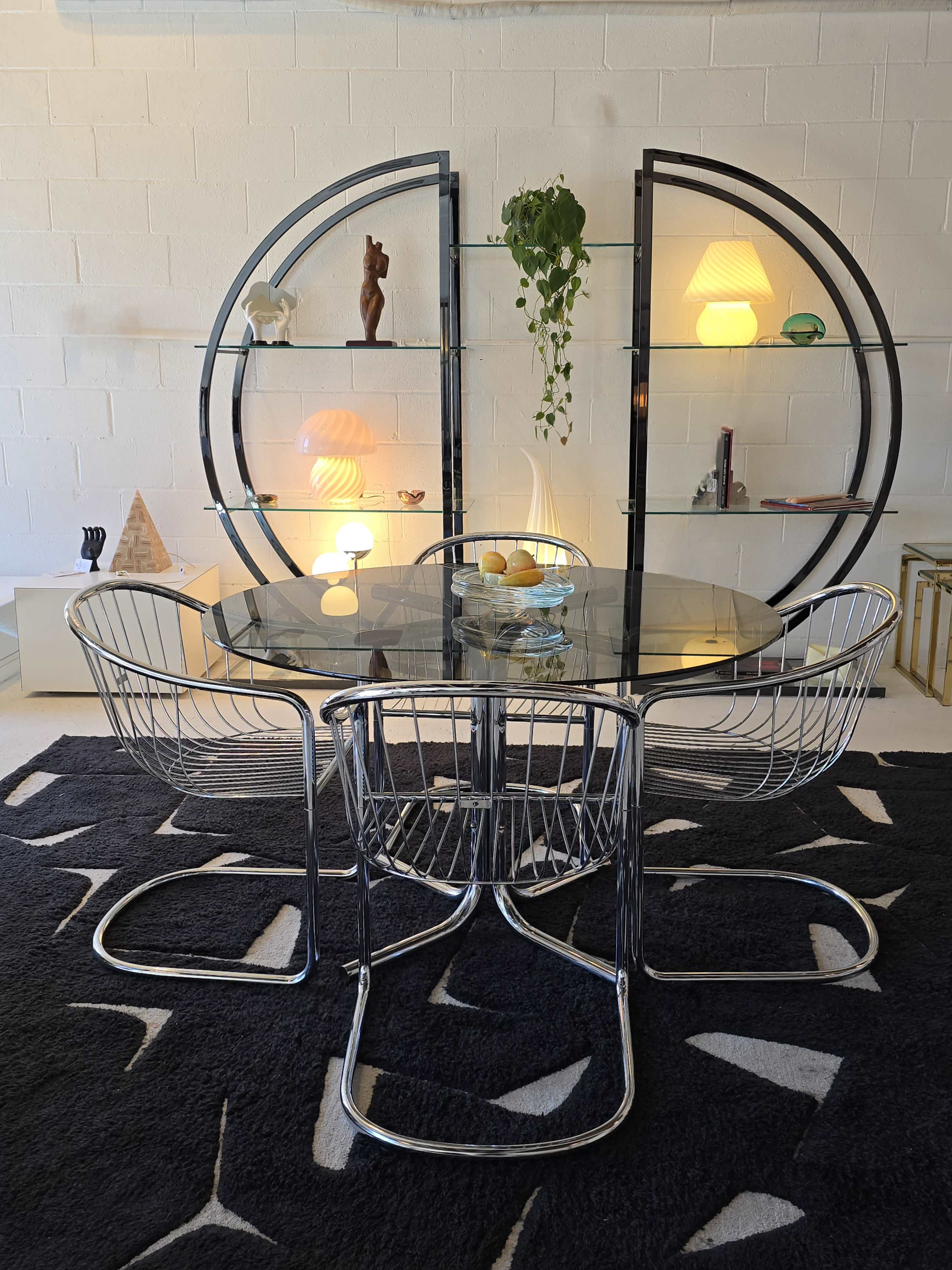 A round glass dining table with wire chairs is set against a backdrop of a modern shelving unit displaying various decorative items and a plant.