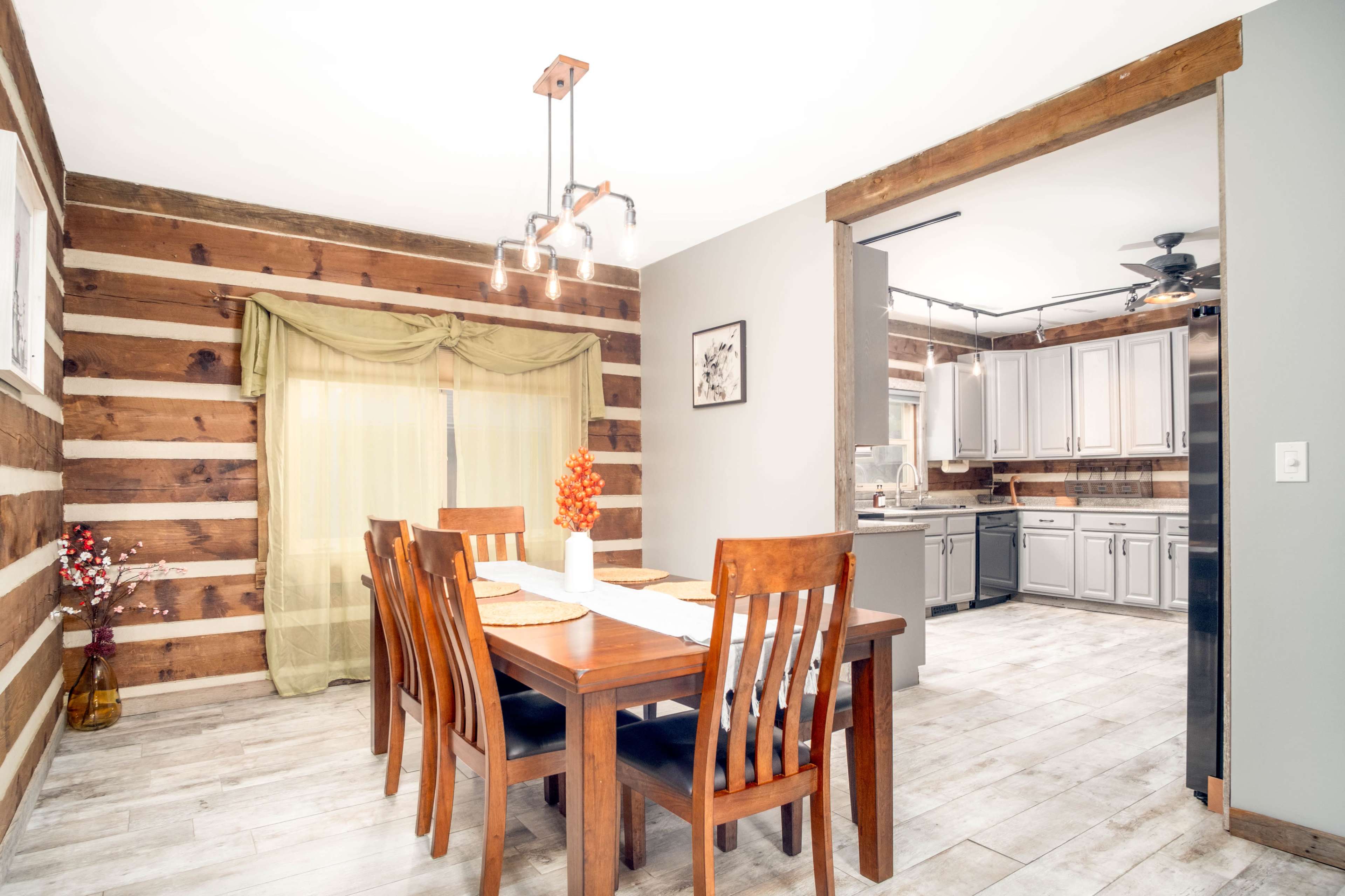 A dining area with a wooden table surrounded by chairs, adjacent to a kitchen featuring grey cabinetry and a rustic log wall design.