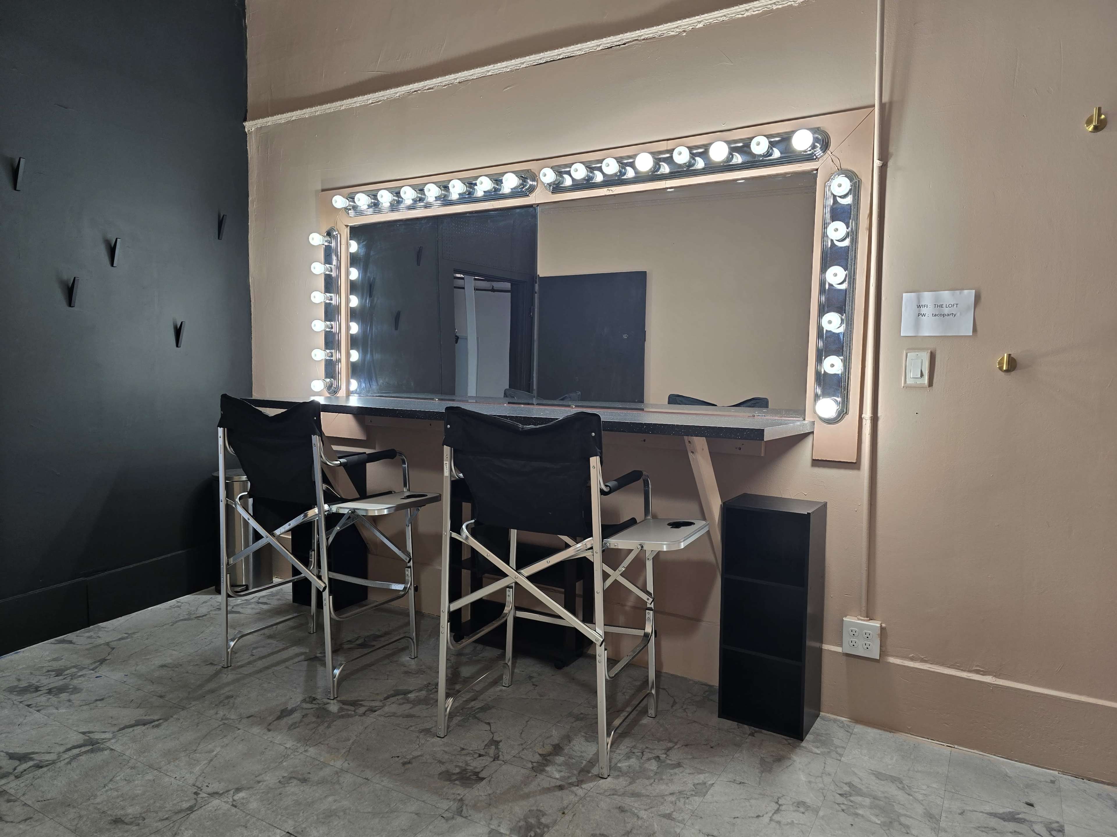 A well-lit vanity area with a large mirror surrounded by bright lights, two black director's chairs, and a small storage unit nearby.