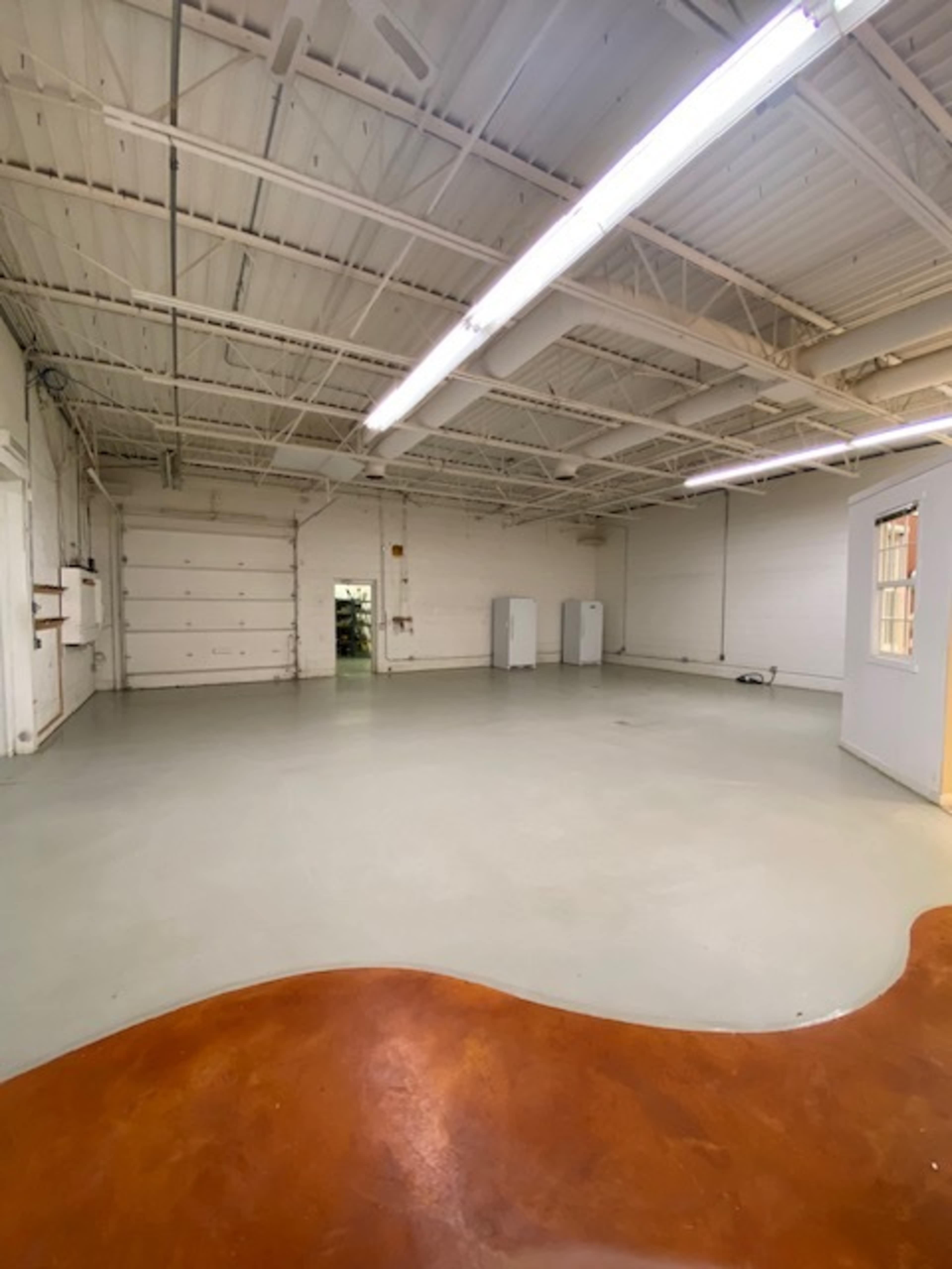A spacious, empty room with a smooth floor and white walls, featuring overhead lighting and a few appliances against one wall.