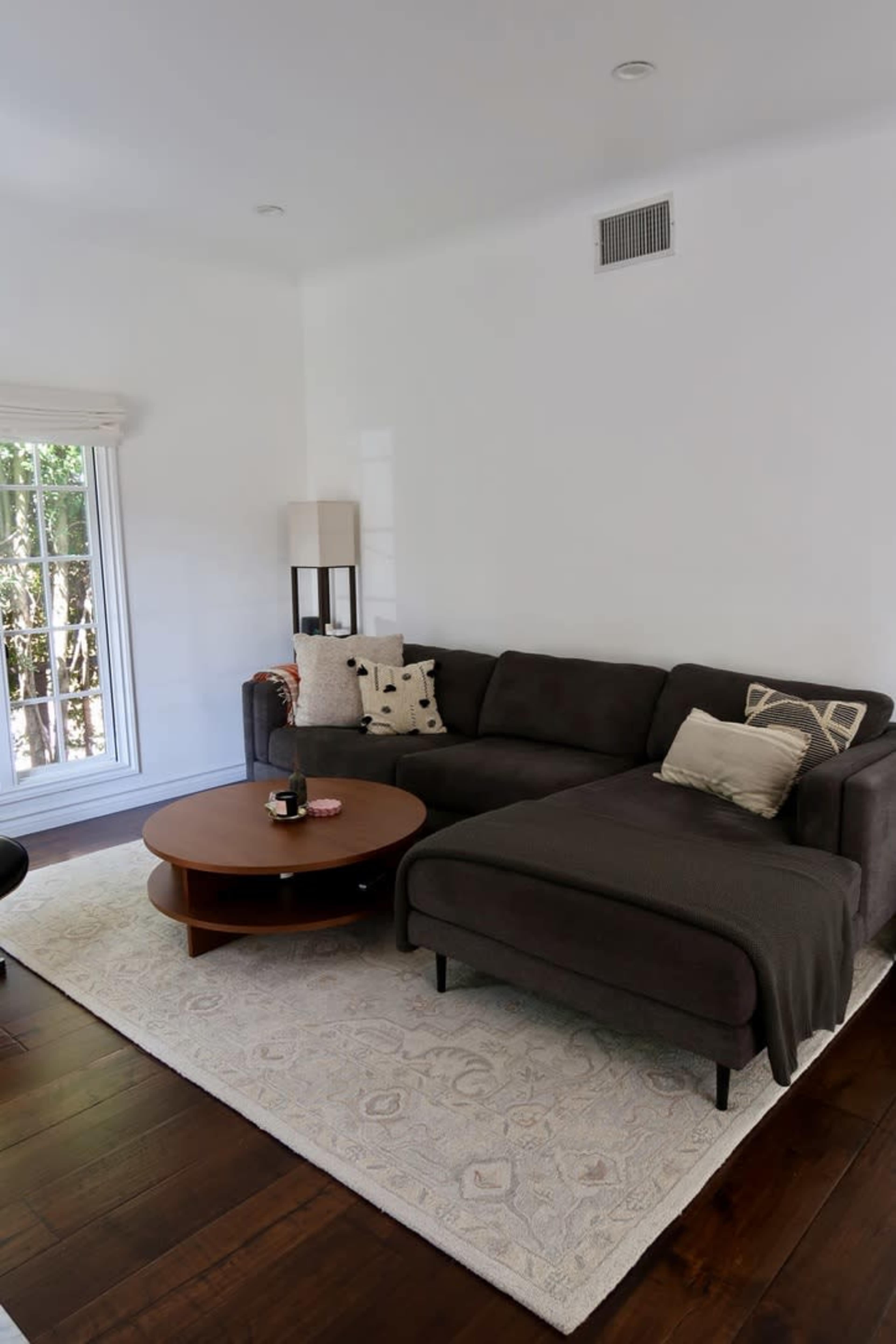 A modern living room features a dark sectional sofa, a round wooden coffee table, and a large area rug on polished wooden floors, with natural light coming through a window.