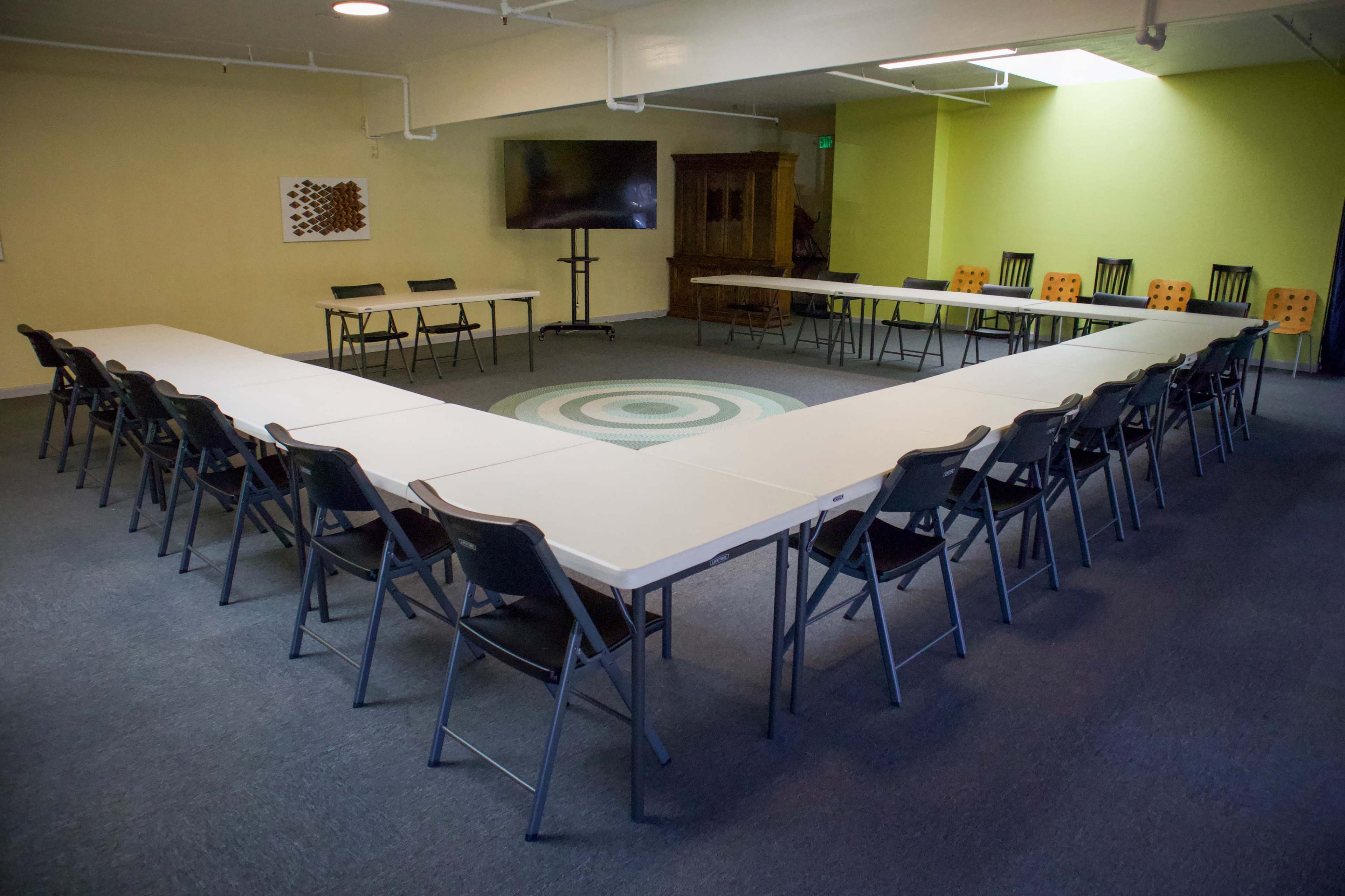 A large meeting room features a U-shaped arrangement of tables and chairs, with a central circular rug and a television screen on one wall.