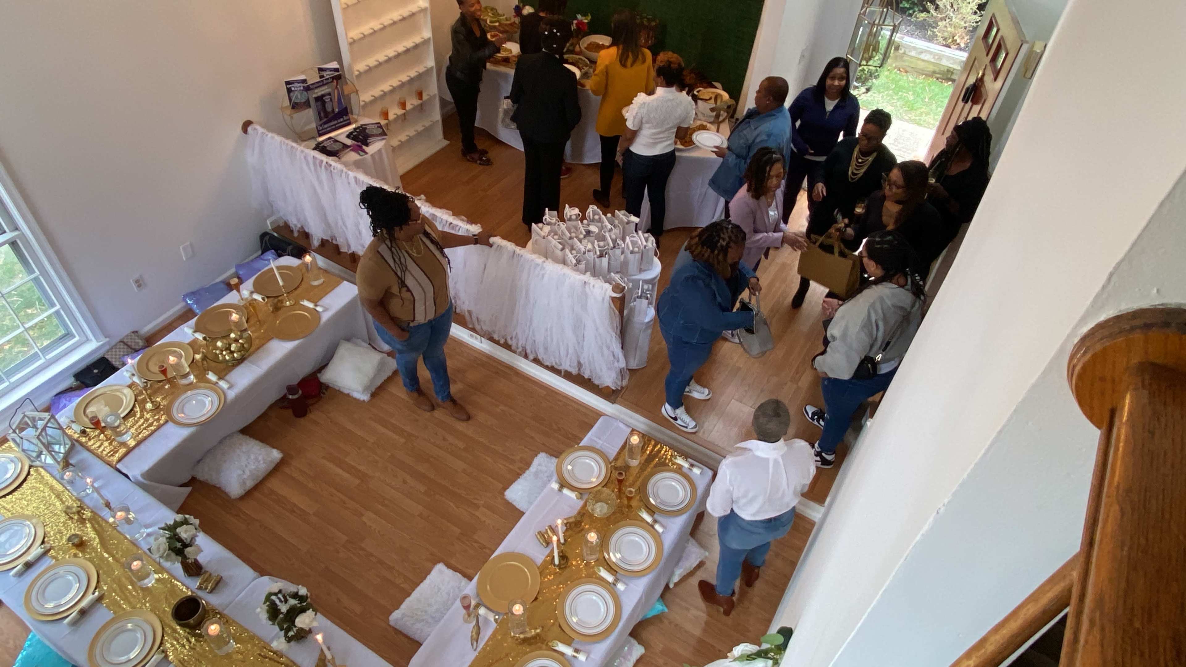 A gathering is taking place in a bright interior space, where people are mingling around tables set with gold and white decorations.