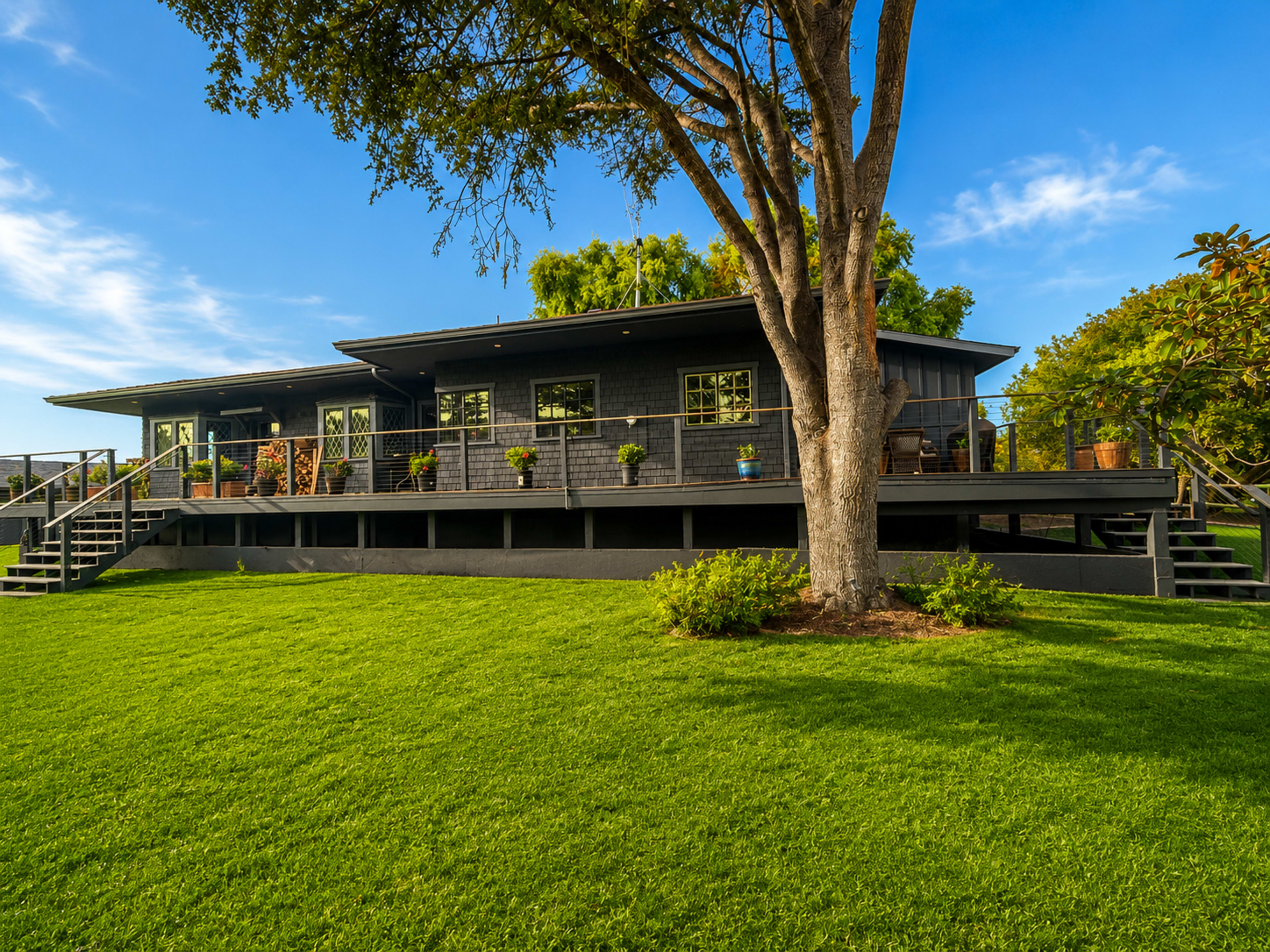 A modern black house with a large deck and potted plants sits on a lush green lawn beneath a blue sky.