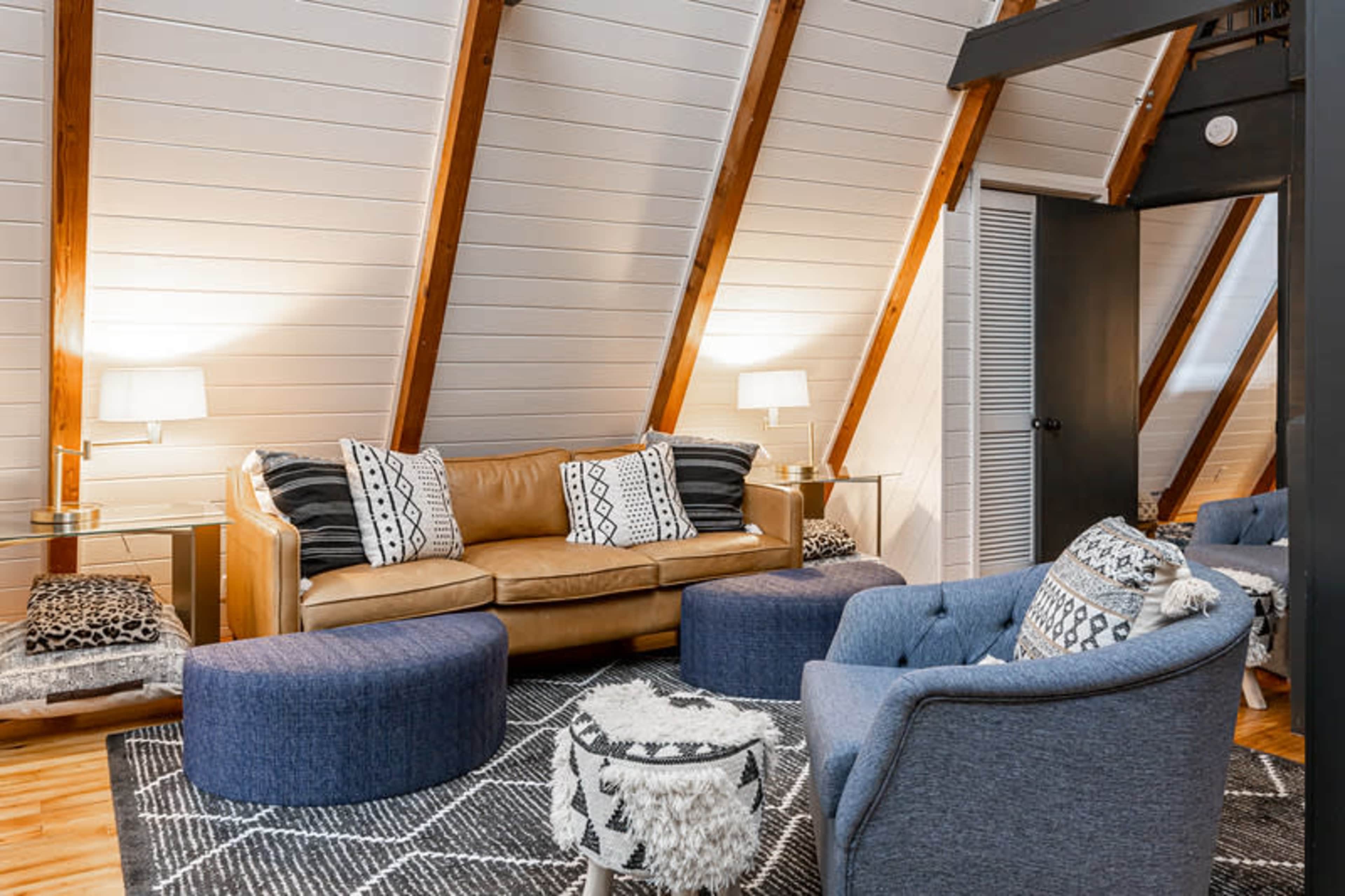 The image shows a cozy living room with sloped wooden ceilings, featuring a brown leather sofa, two blue ottomans, and a round coffee table on a patterned rug.