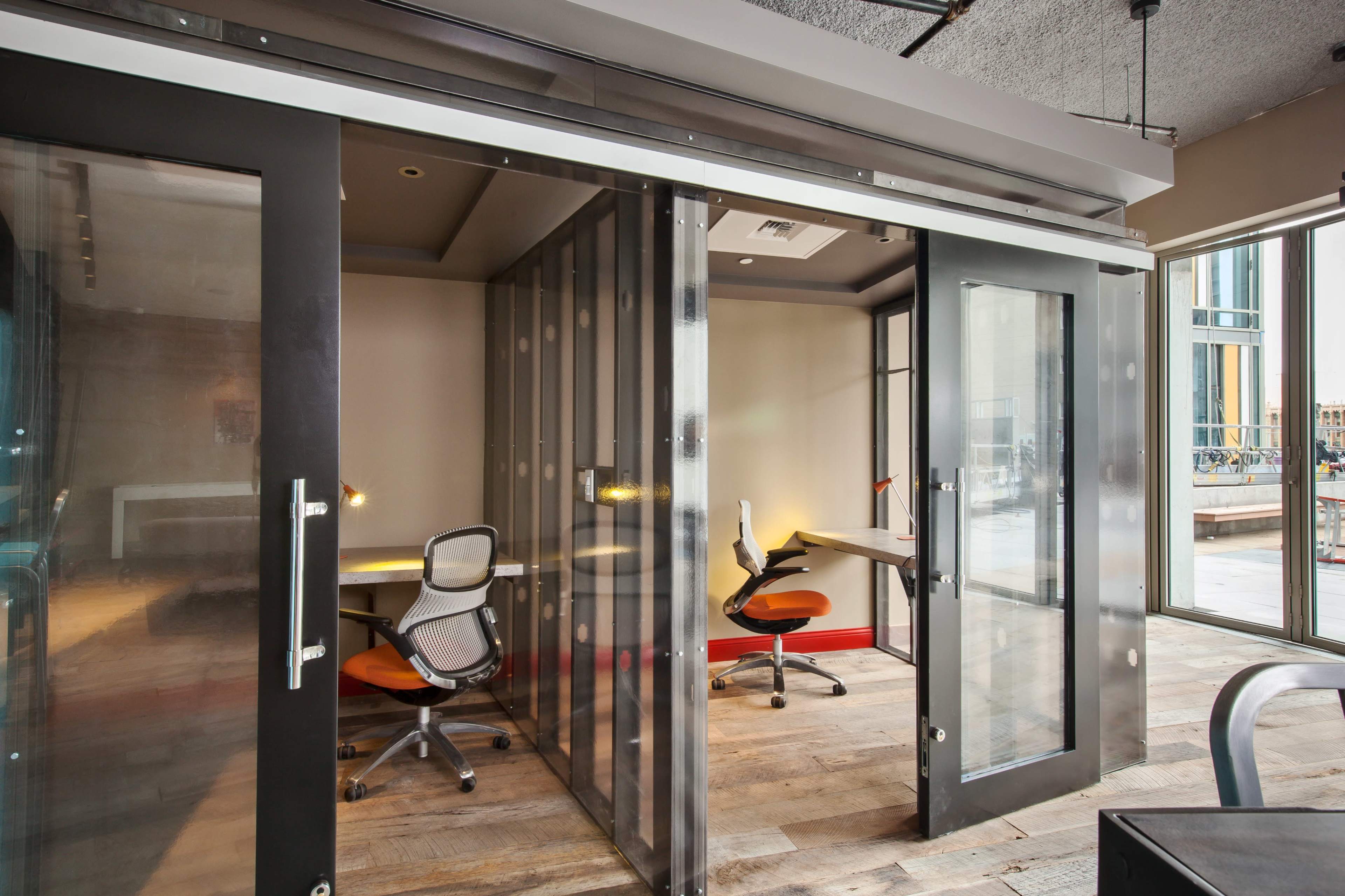 A modern office space featuring glass-walled meeting rooms with desks and rolling chairs.