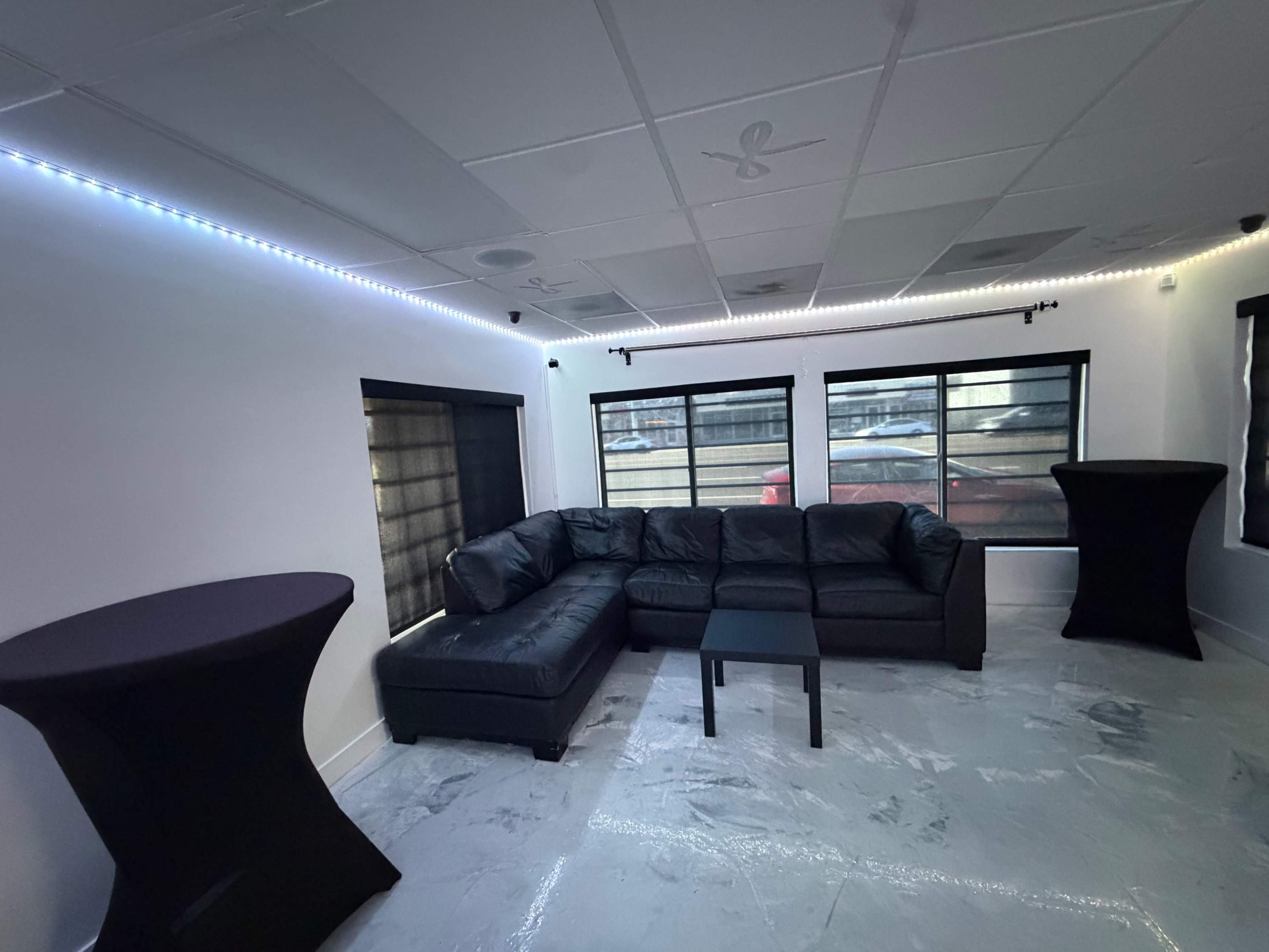 The image shows a modern room with a black sectional sofa, a small black table, and two tall black cocktail tables, illuminated by LED lighting along the walls and featuring large windows.
