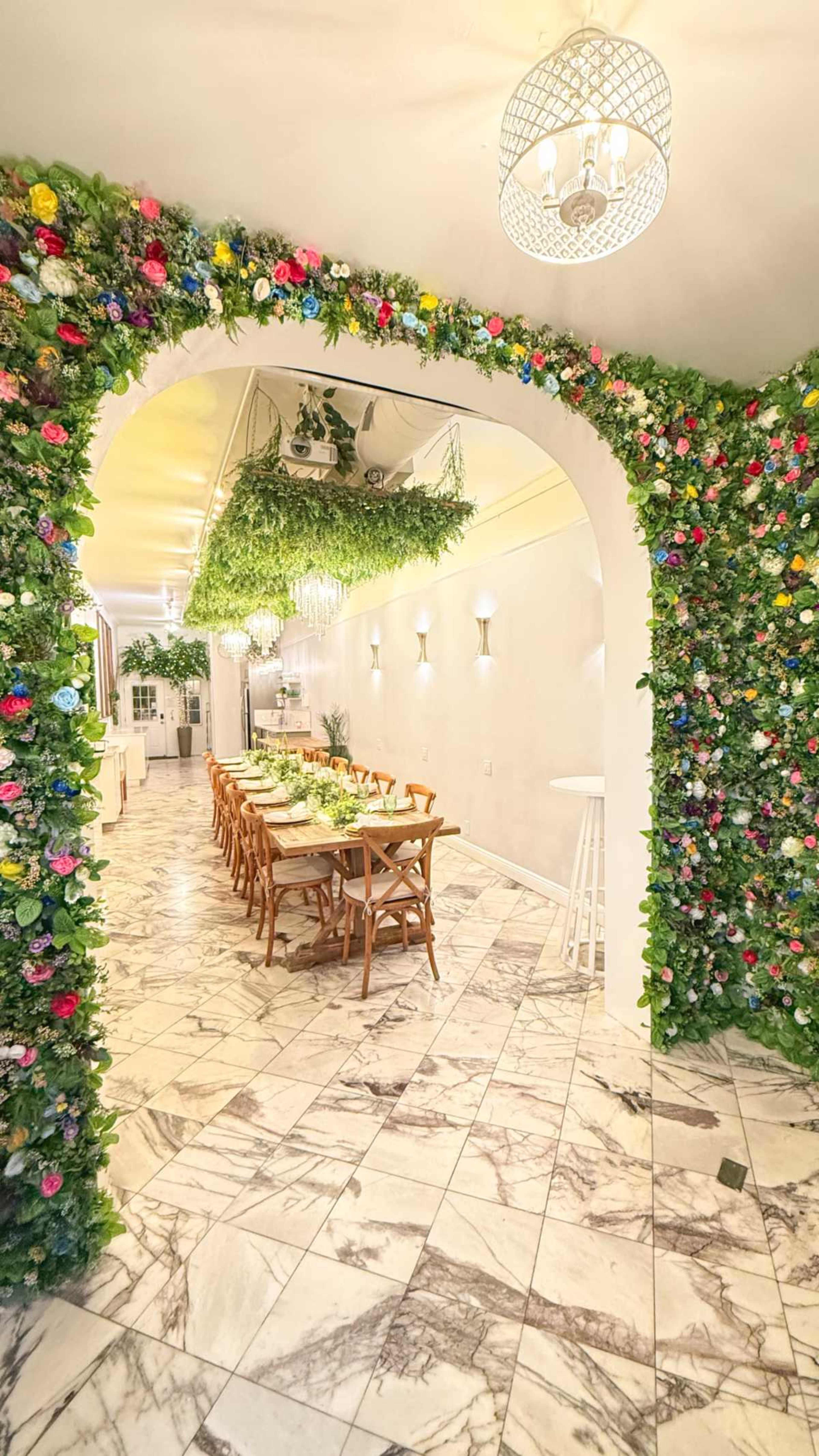 A floral archway leads into a dining area with a long wooden table, surrounded by green foliage and flower decorations, set against a marble floor.