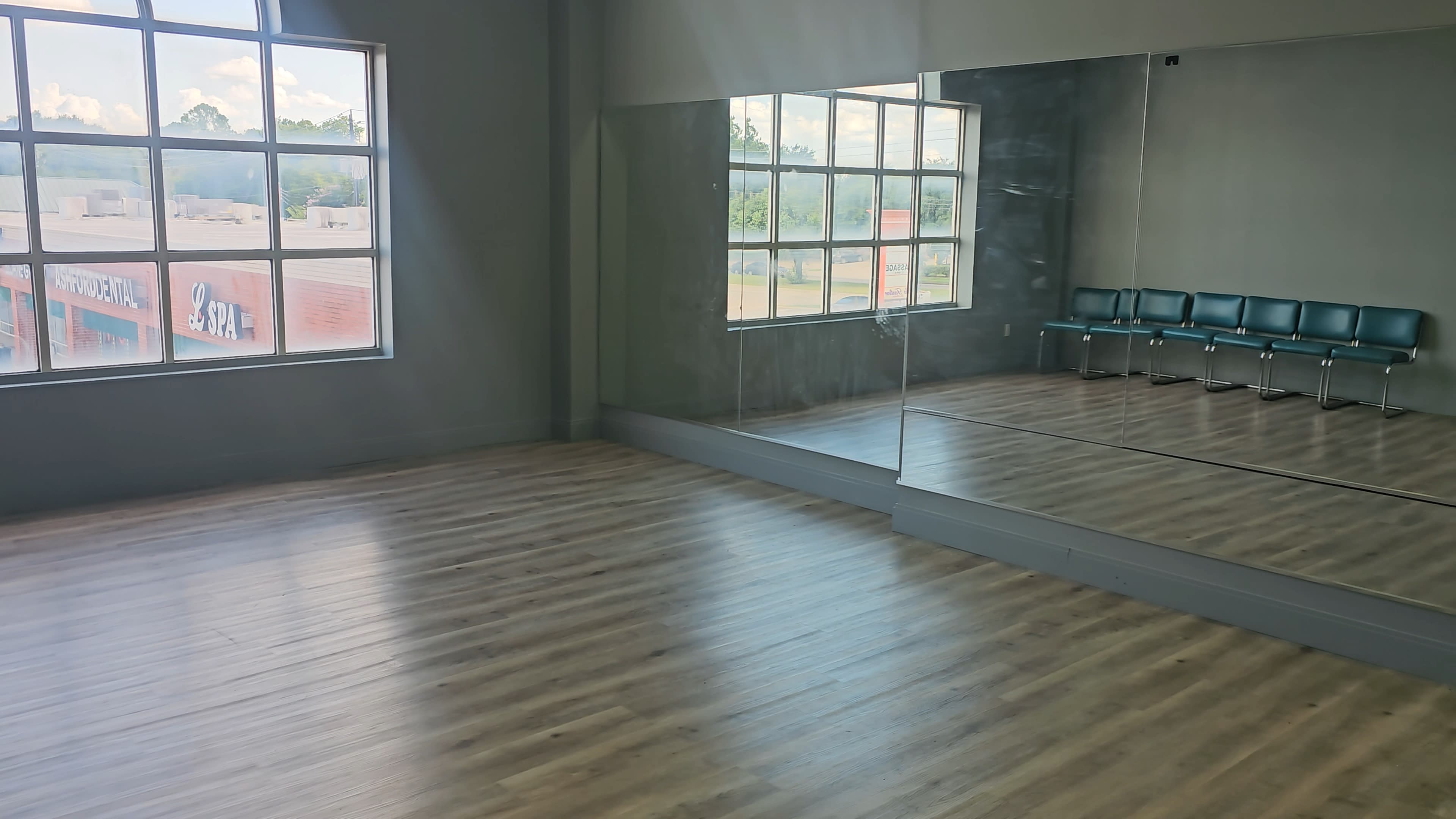 A spacious studio with a wooden floor, large windows, and a mirrored wall, accompanied by a row of teal chairs.