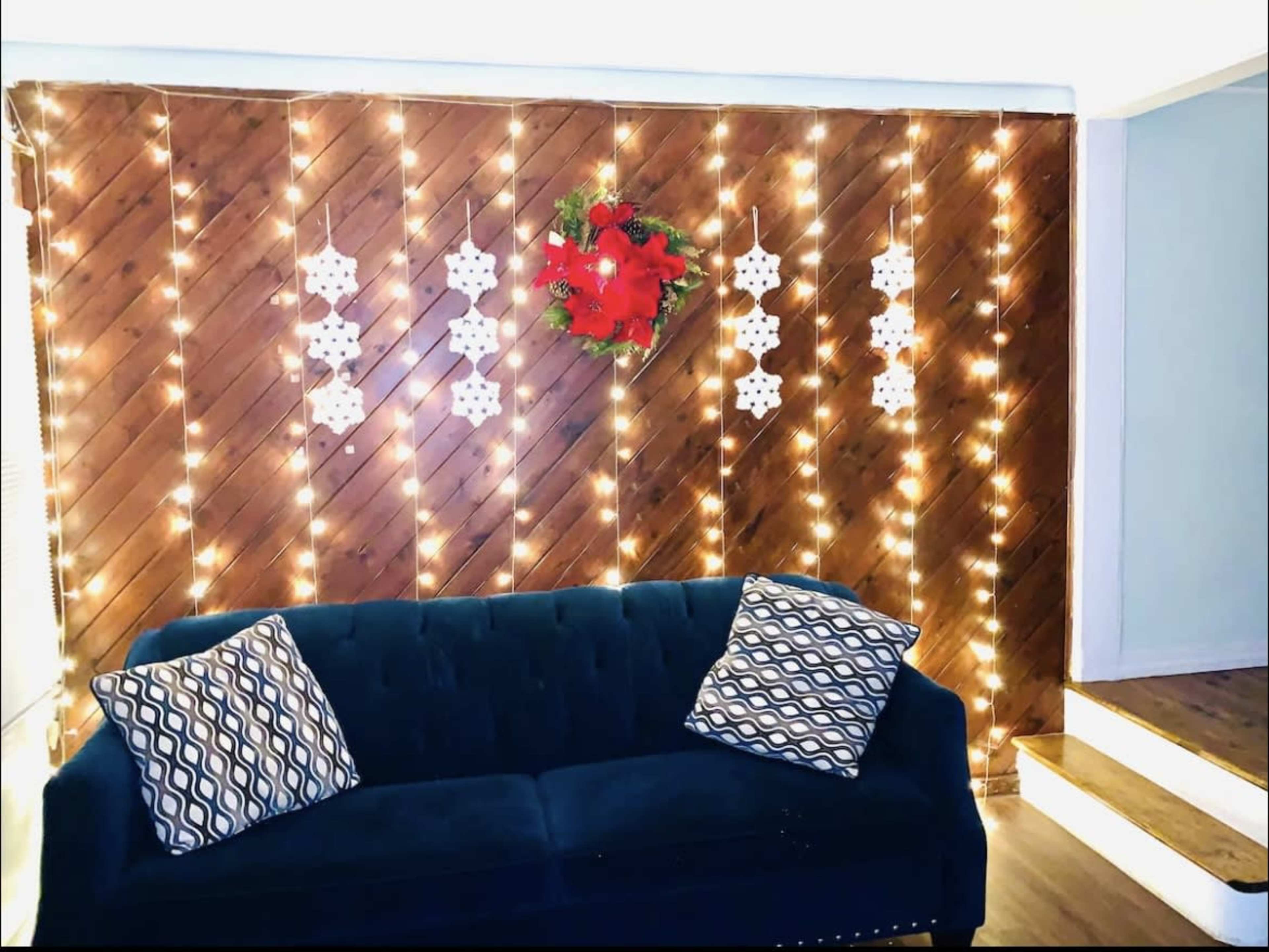 A blue couch with patterned pillows sits against a wooden wall adorned with string lights and decorative snowflakes, complemented by a festive wreath.