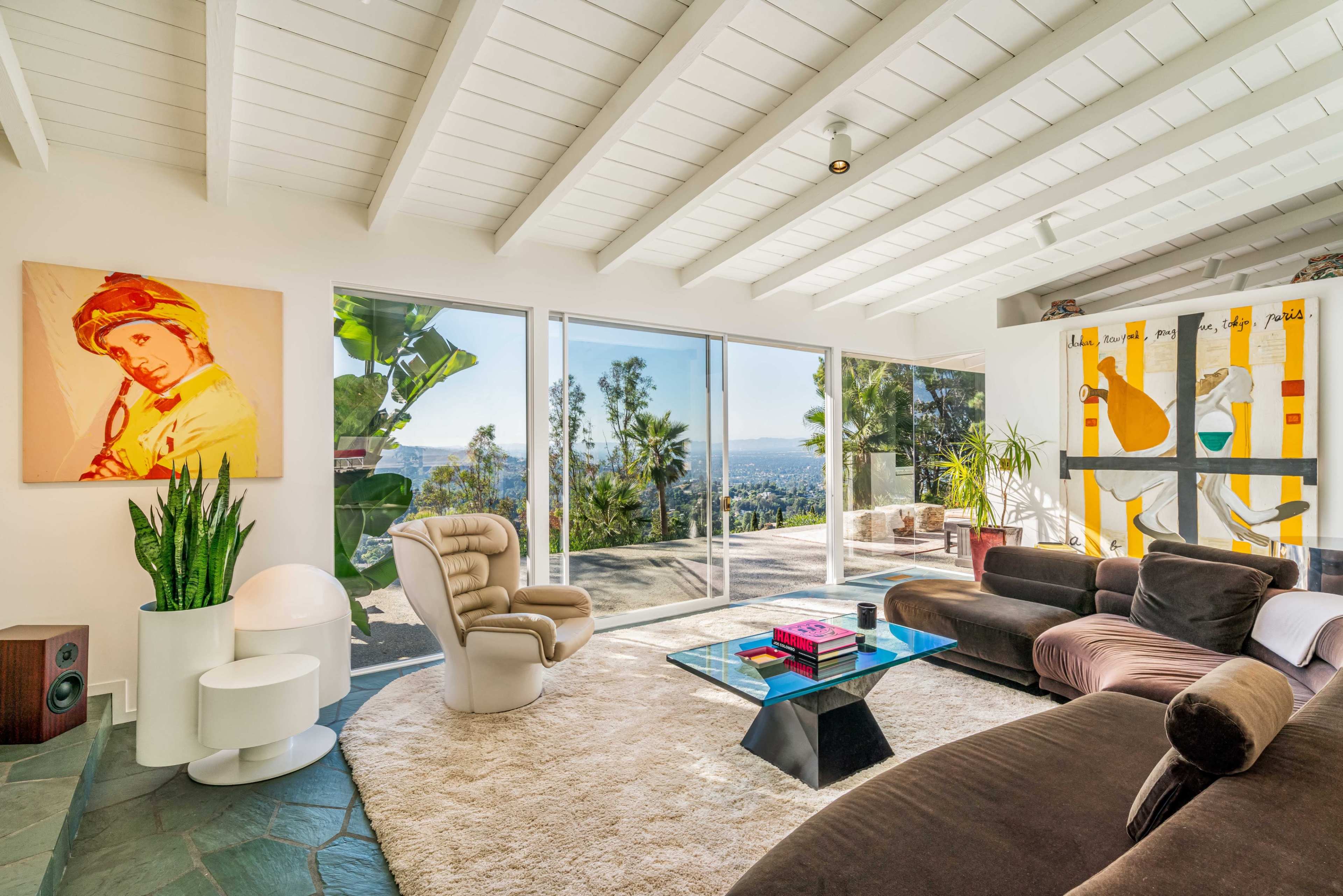 A spacious living room features large glass windows overlooking a scenic view, modern furniture, and vibrant artwork on the walls.