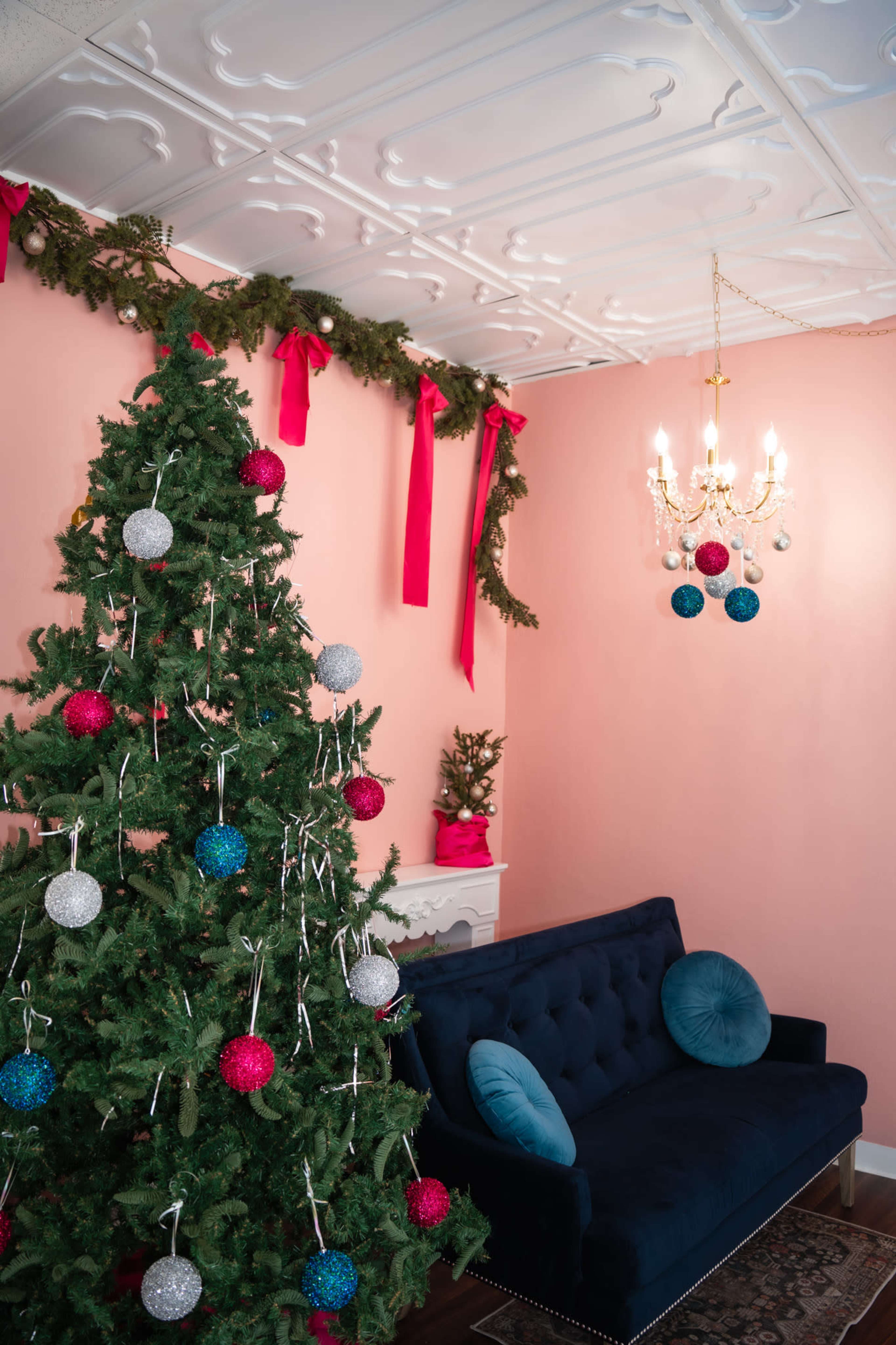 A decorated Christmas tree stands next to a blue sofa in a room with pink walls and holiday garlands.