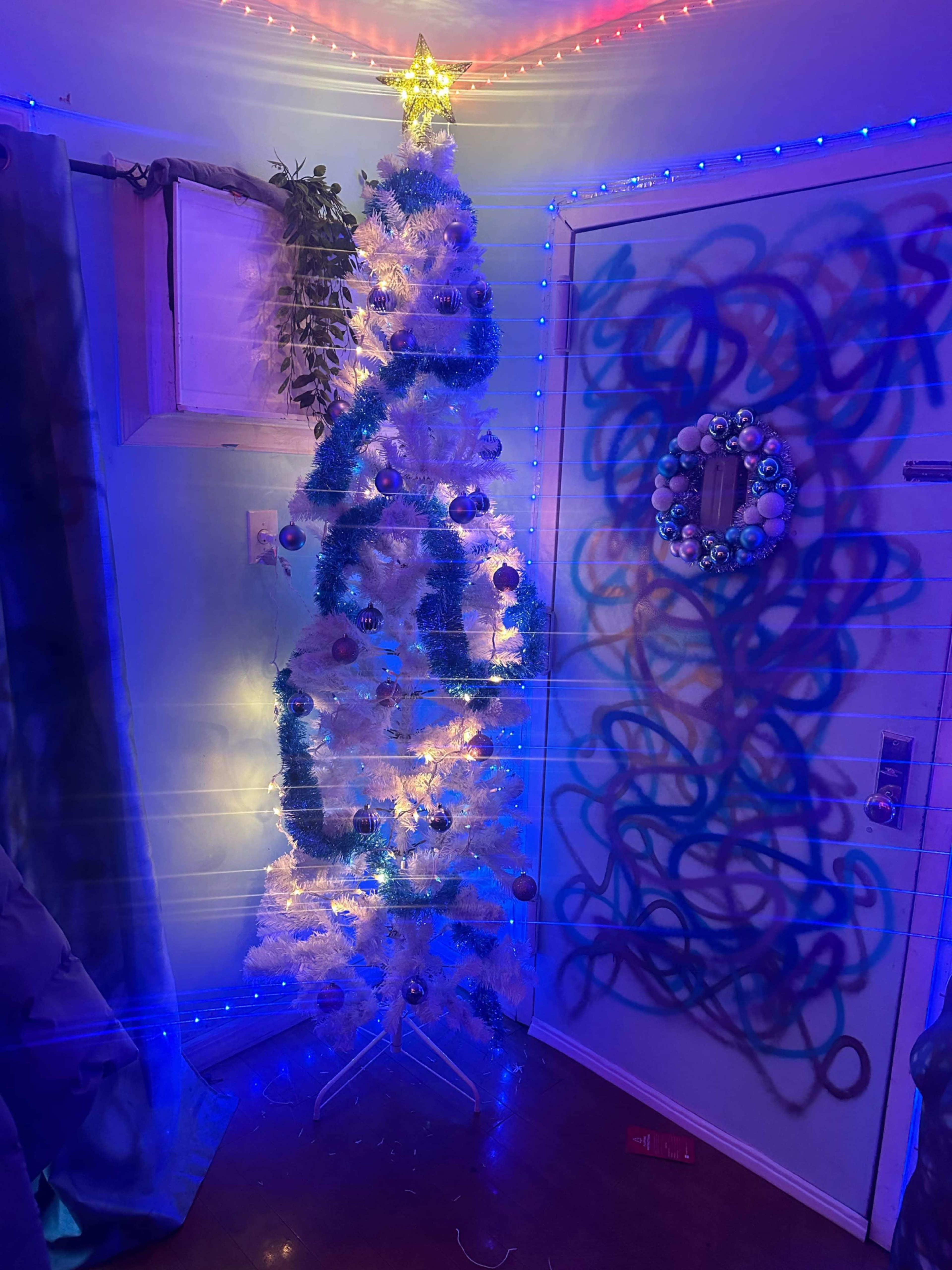 A Christmas tree decorated with lights and ornaments stands in a room illuminated by colored lighting.