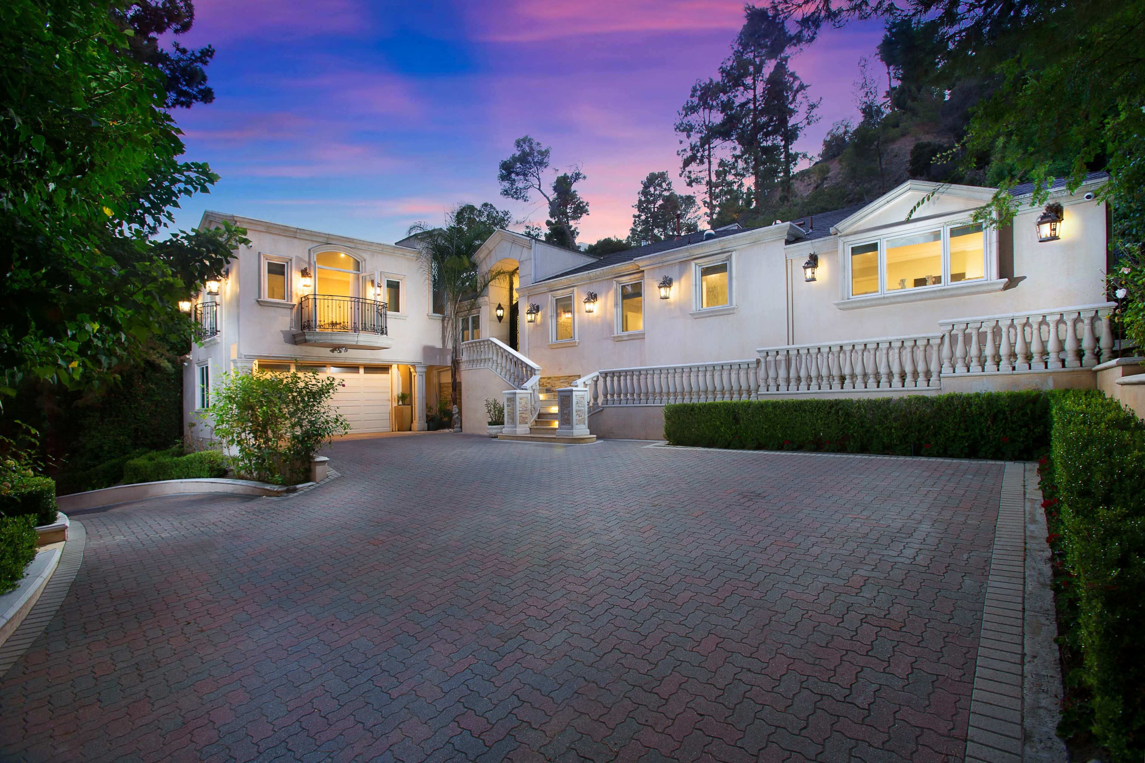 A spacious mansion with a well-paved driveway, surrounded by trees and illuminated by exterior lights during twilight.