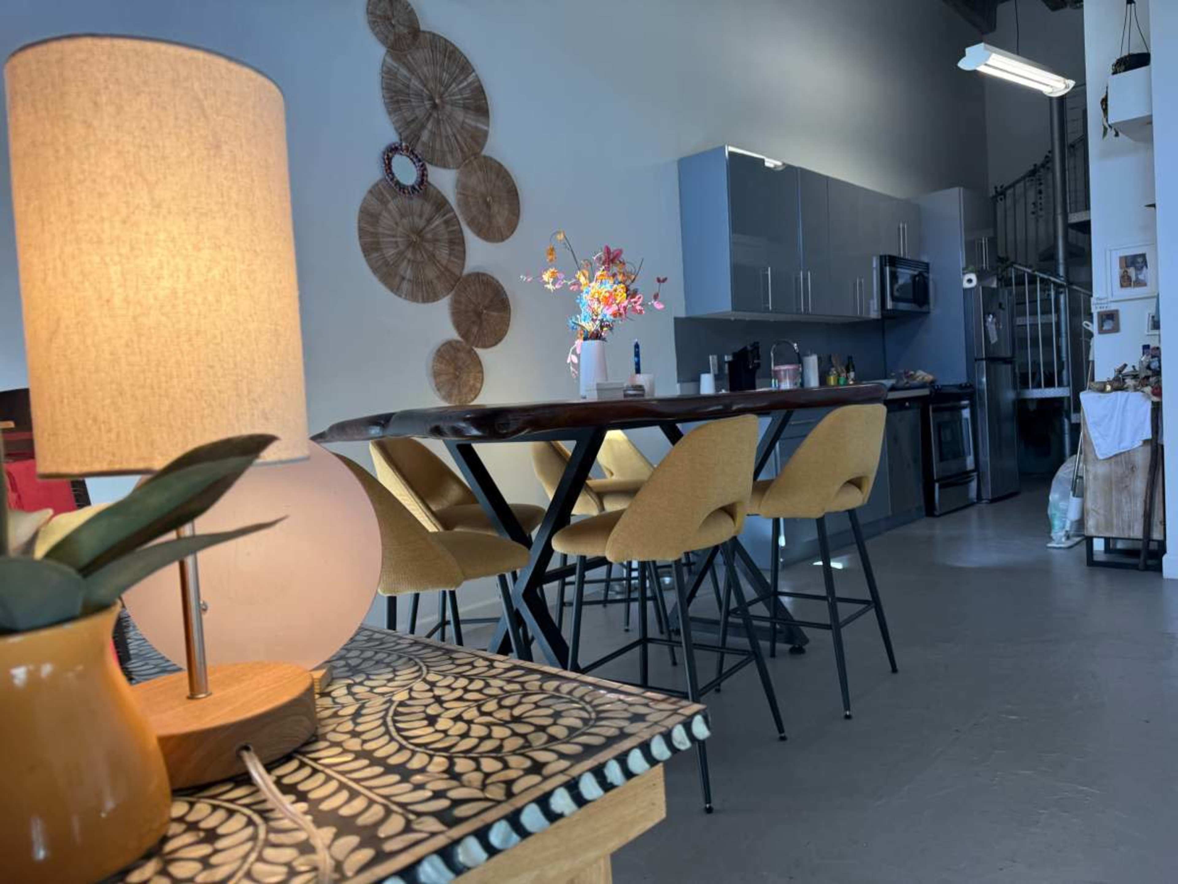 The image shows a modern kitchen and dining area with a large wooden table surrounded by yellow chairs, alongside a decorative lamp and a colorful floral arrangement.